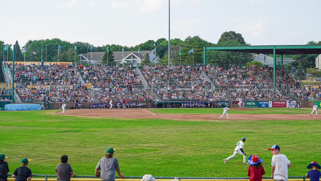 A baseball game in progress at a stadium with a large crowd in the stands. Players are seen on the field, and spectators watch from the stands and behind a fence.