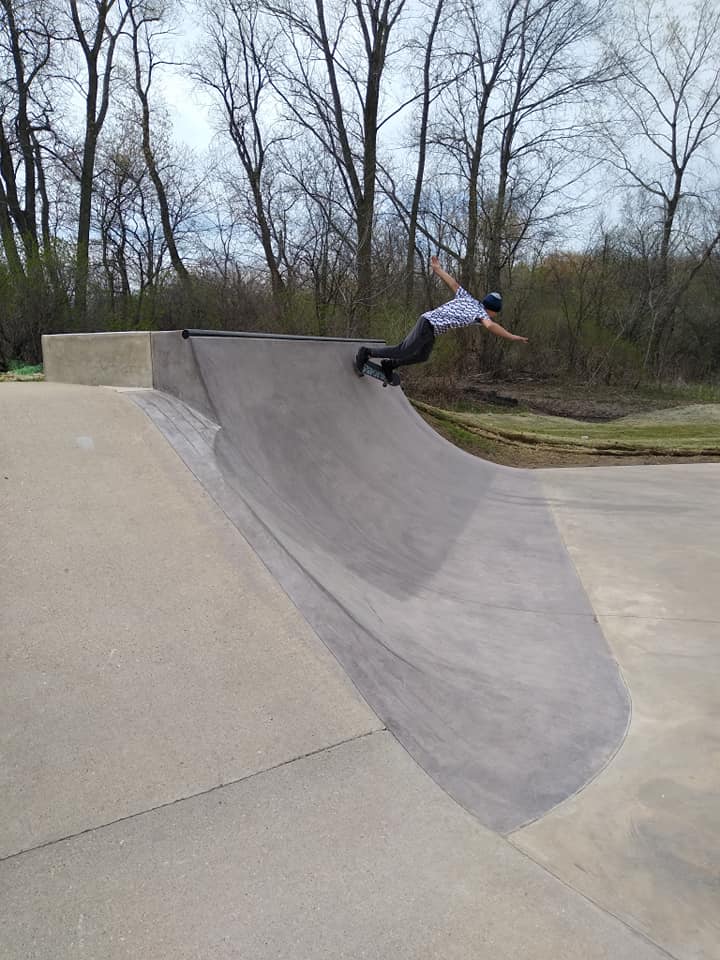 A person wearing a helmet and rollerblades rides along an inclined concrete ramp at an outdoor skatepark with bare trees in the background.