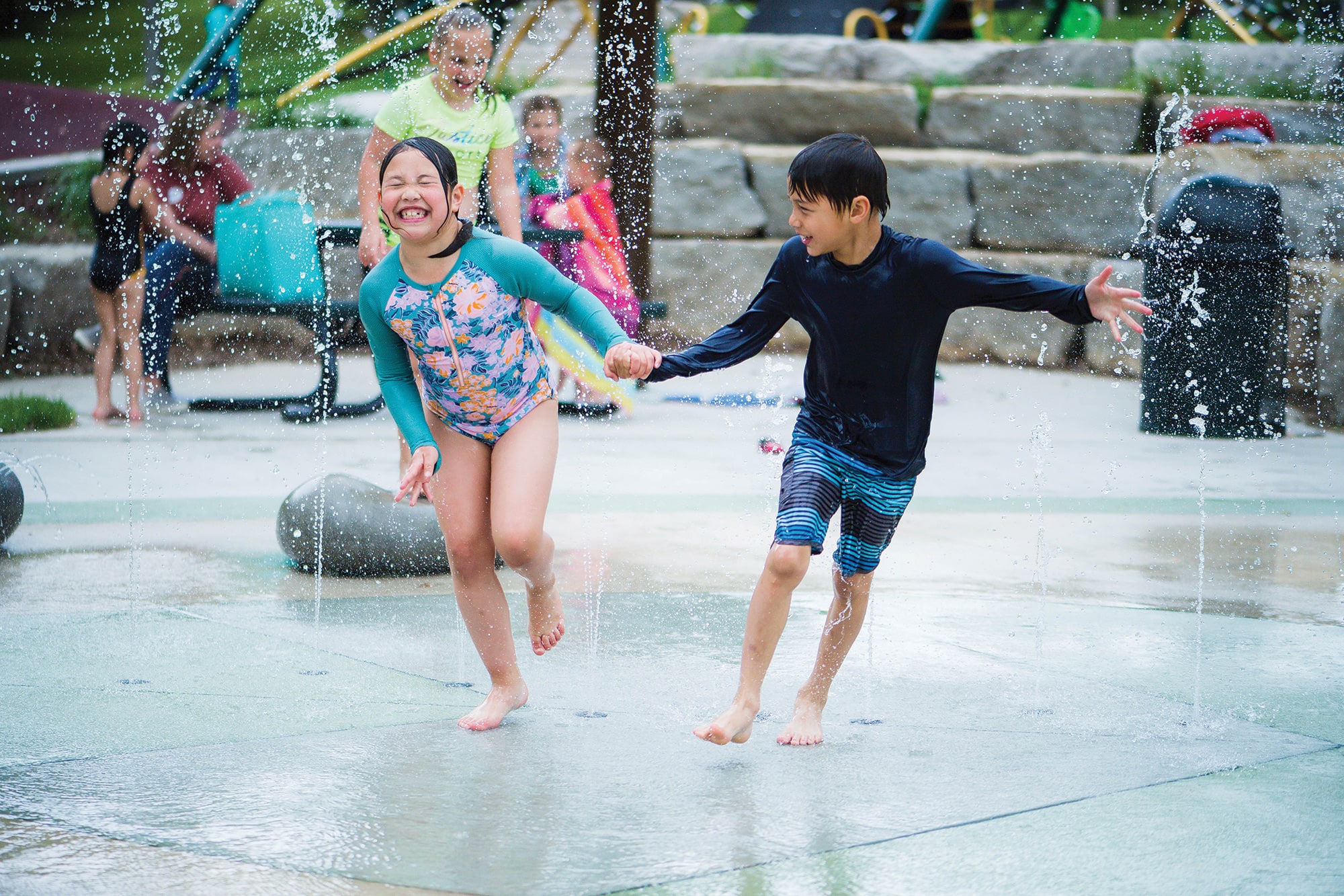 Two children in swimwear run through a splash pad, holding hands and laughing, with water spraying around them. Other children and adults can be seen in the background.