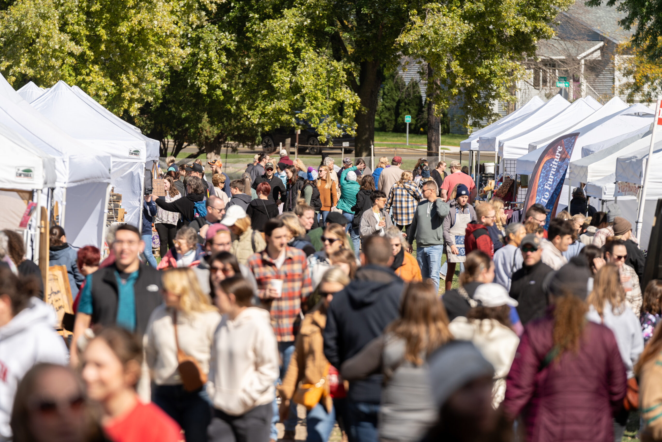 A bustling outdoor market with numerous white tents and people walking and shopping on a sunny day. Trees in the background.