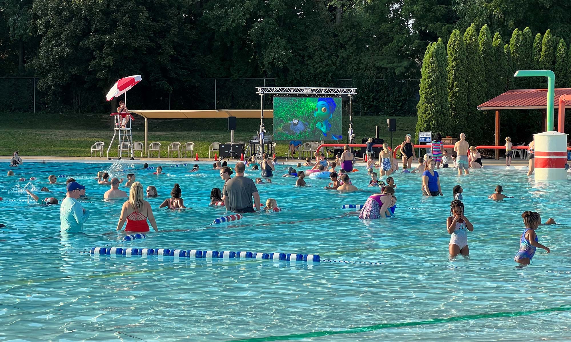 A crowded outdoor swimming pool with people of various ages swimming and playing. A lifeguard watches from a chair, and a large screen displays an animated movie in the background.