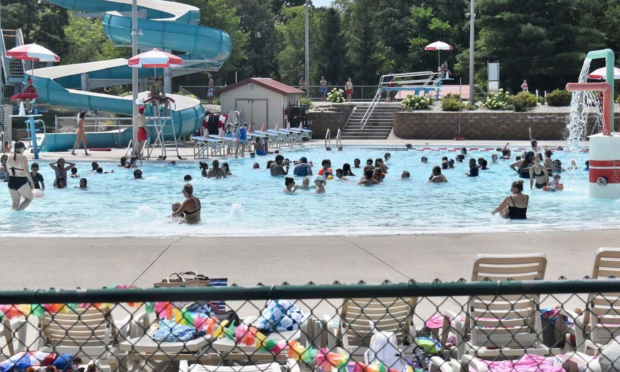 A public swimming pool with many people swimming and playing. A large water slide and a splash feature are present. The poolside area has chairs and towels, with a fence in the foreground.