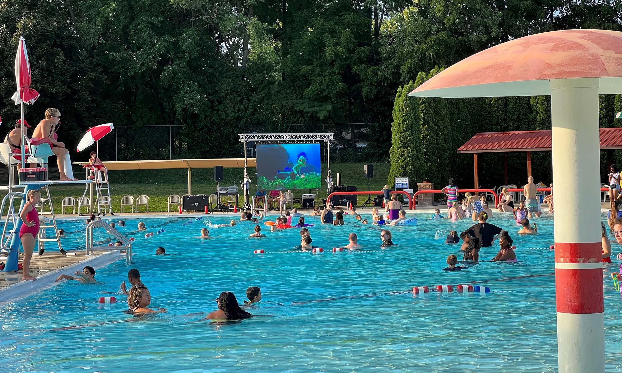 A public swimming pool with many people swimming and socializing; a large screen displays a movie, and lifeguards oversee the activity.