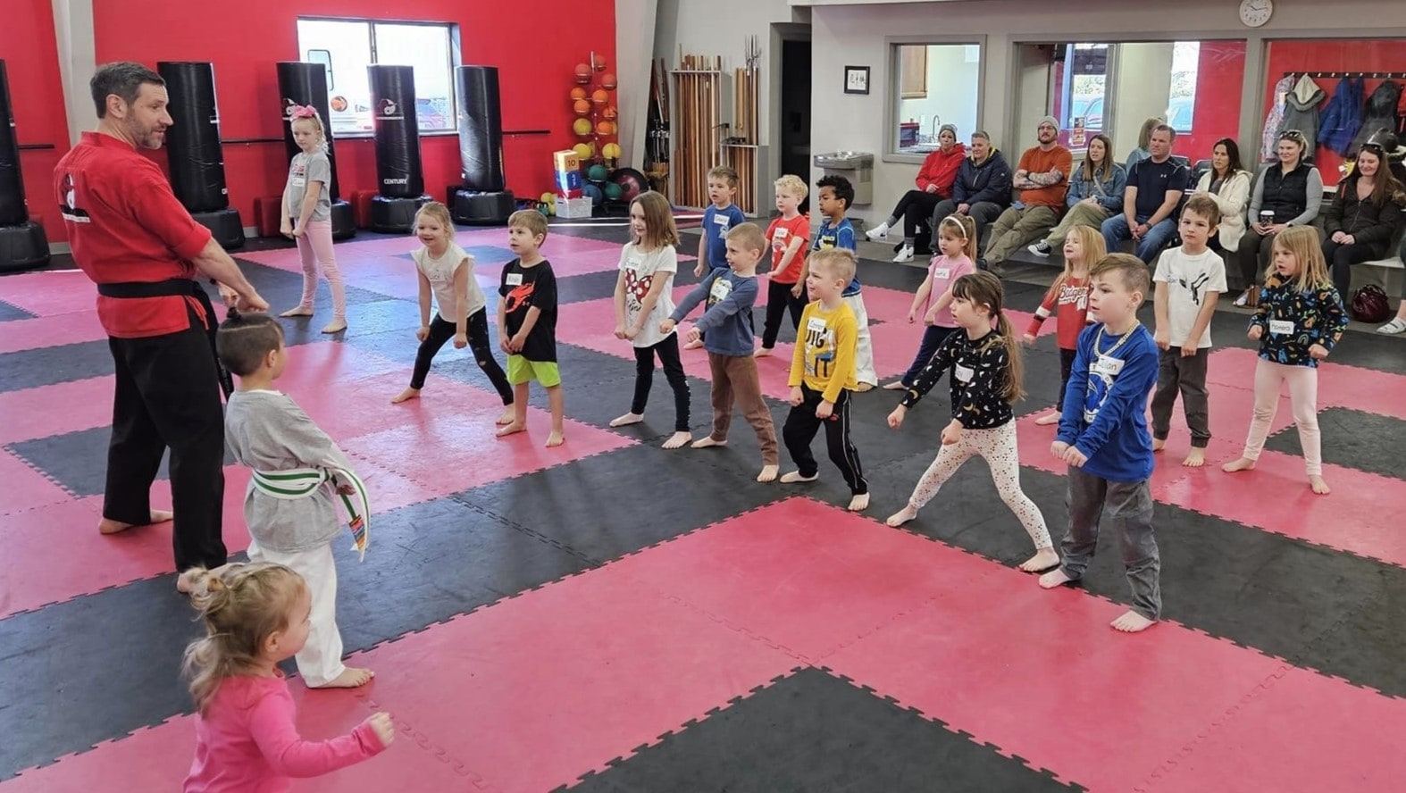 A group of young children in a martial arts class stand in formation on a mat as an instructor in a red uniform gives directions. Parents are seated in the background, observing the class.