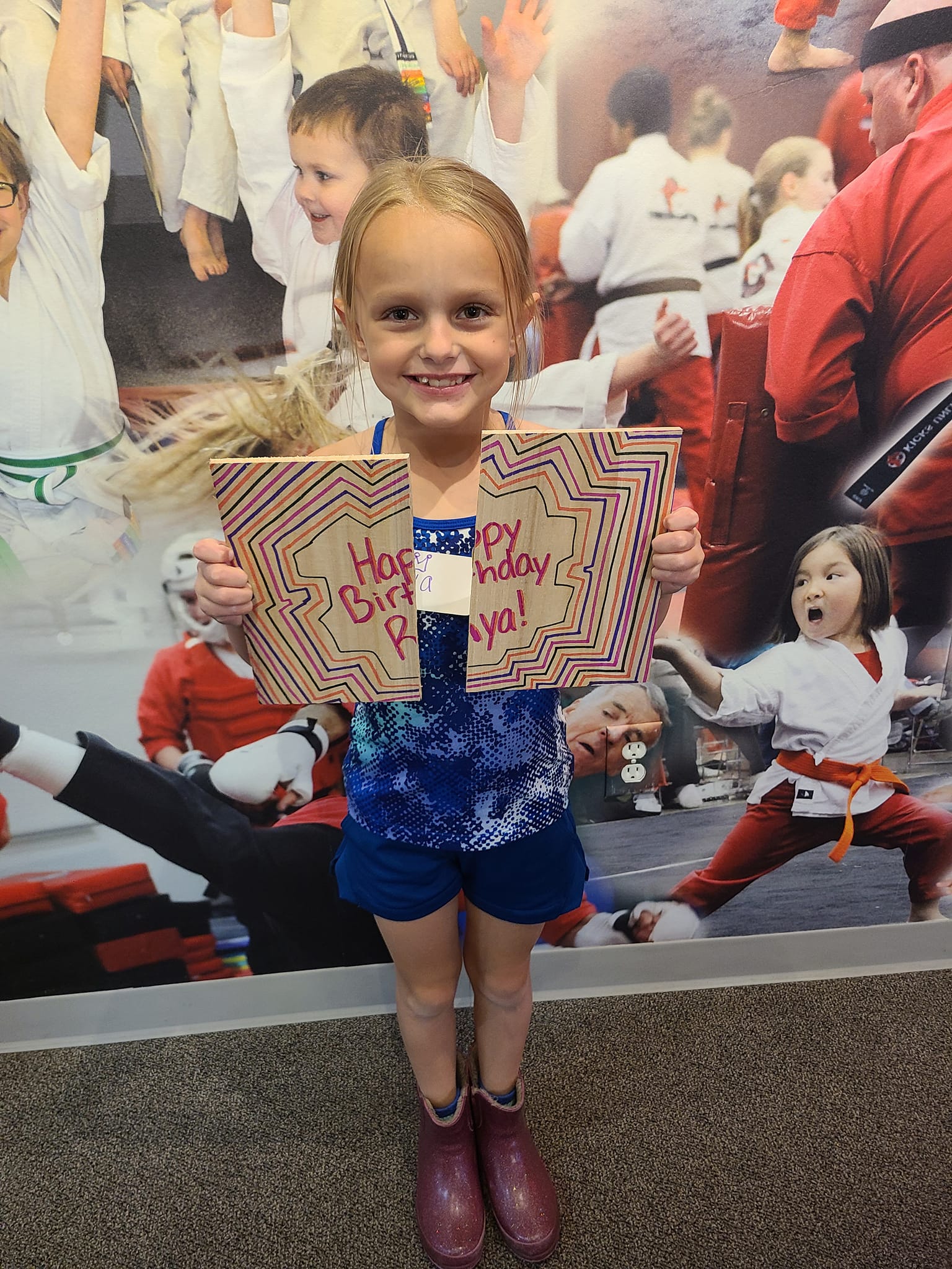 A young girl with blonde hair holding a "Happy Birthday Raya!" card stands in front of a colorful wall with images of children in martial arts uniforms. She is wearing a blue outfit and purple boots.