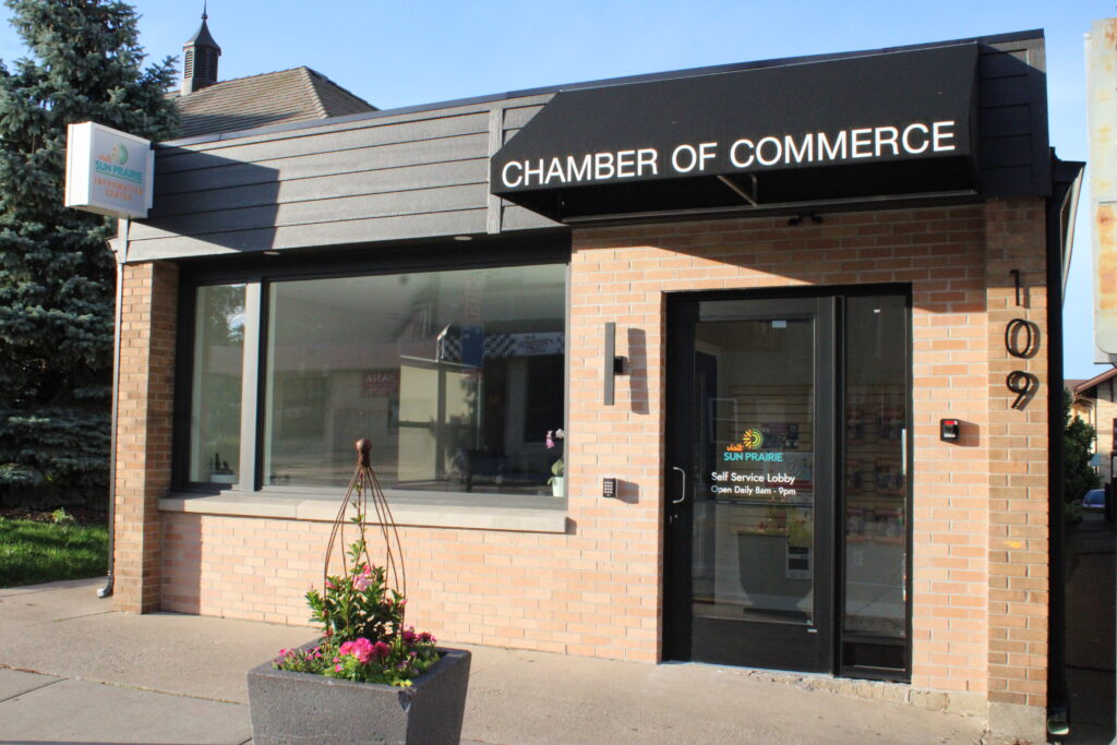 Small brick building labeled "Chamber of Commerce" with a potted plant outside, large window, and glass door displaying business hours.