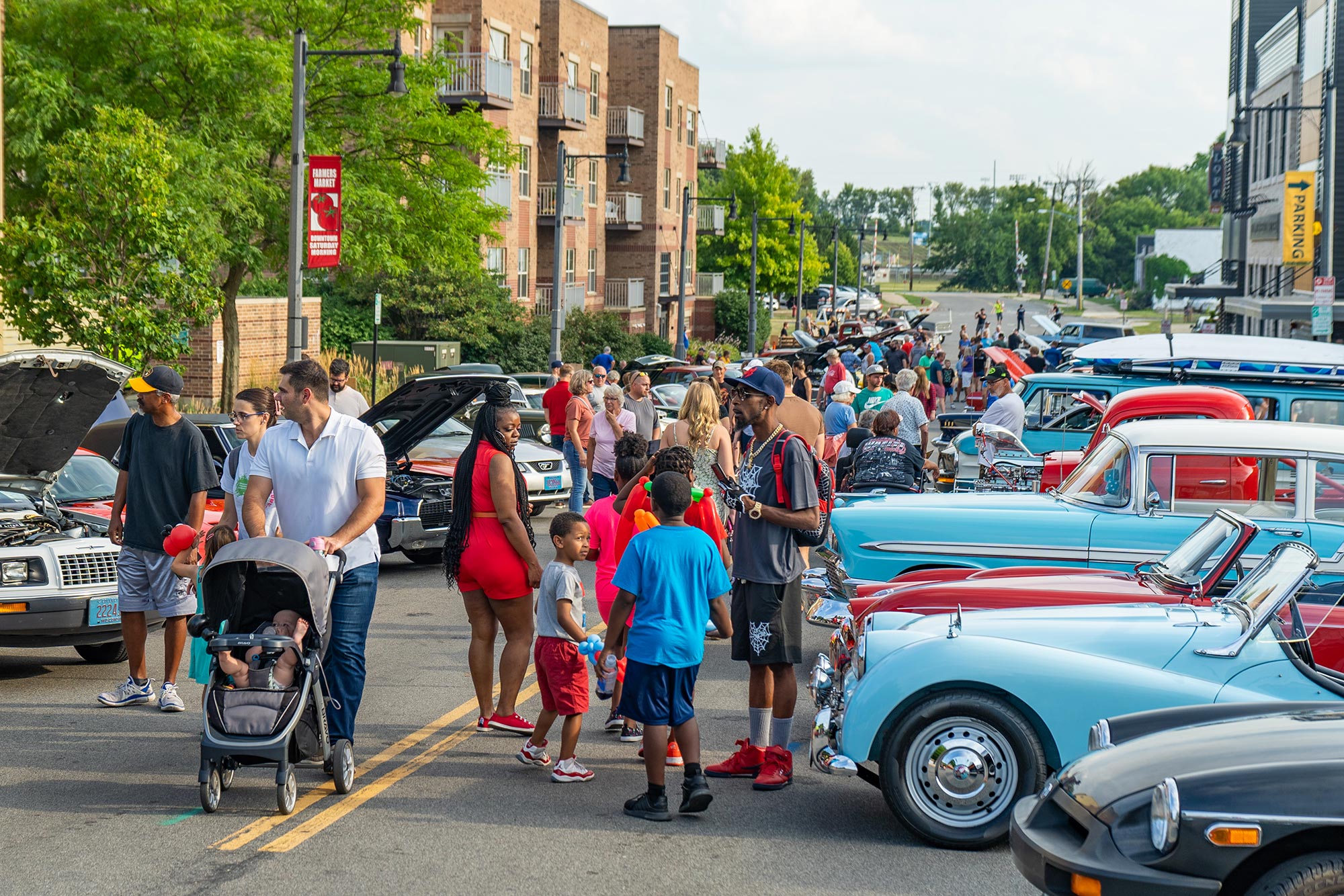 People attending an outdoor car show in a city. Various vintage cars are displayed along a street, while attendees, including families with children, walk around and observe the vehicles.