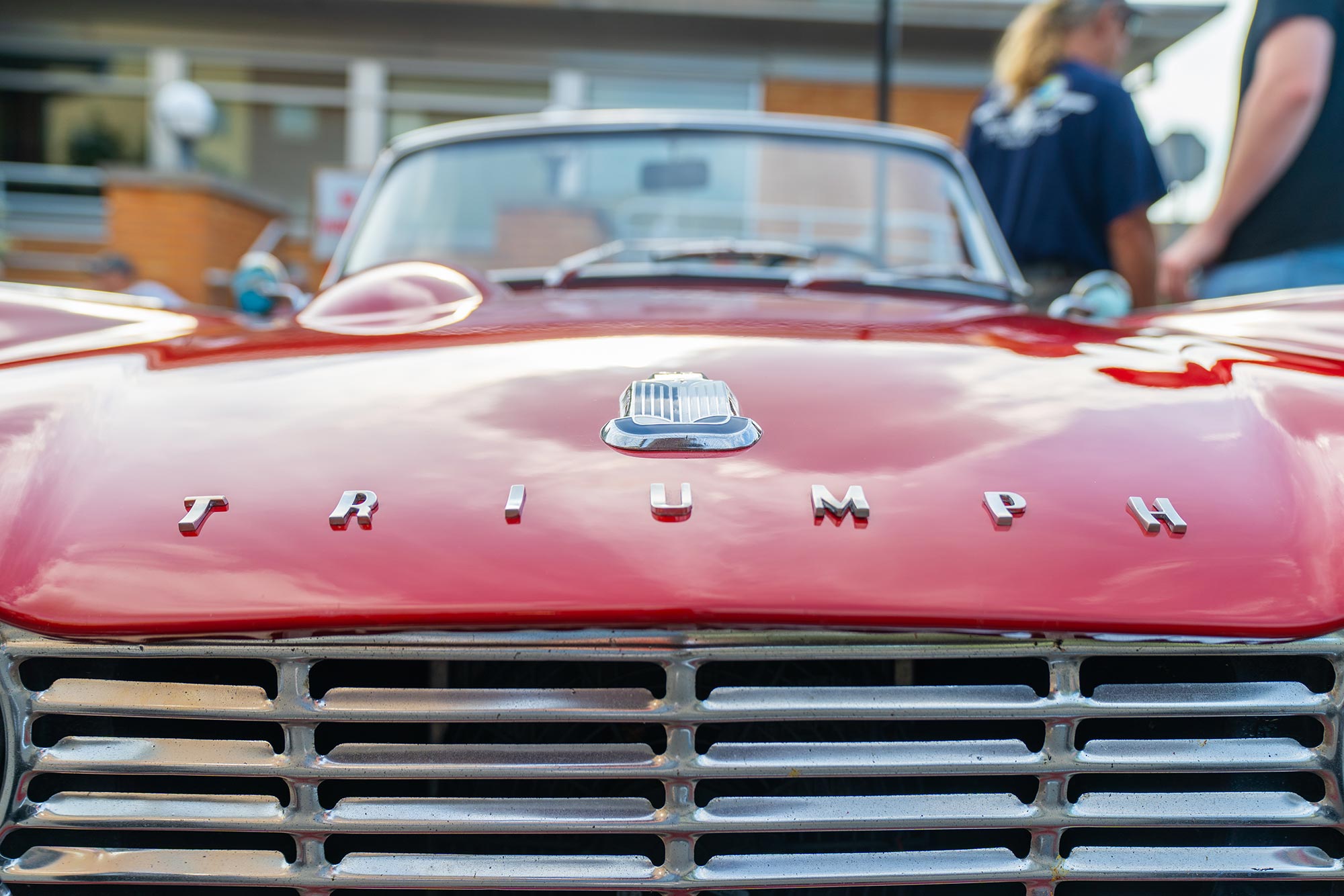 Close-up shot of the front grille and badge of a red Triumph convertible car, with the background slightly blurred.