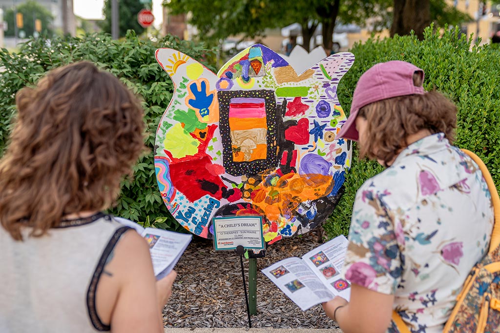 Two people read information from pamphlets in front of a colorful, abstract art display shaped like a flower petal, in an outdoor setting.