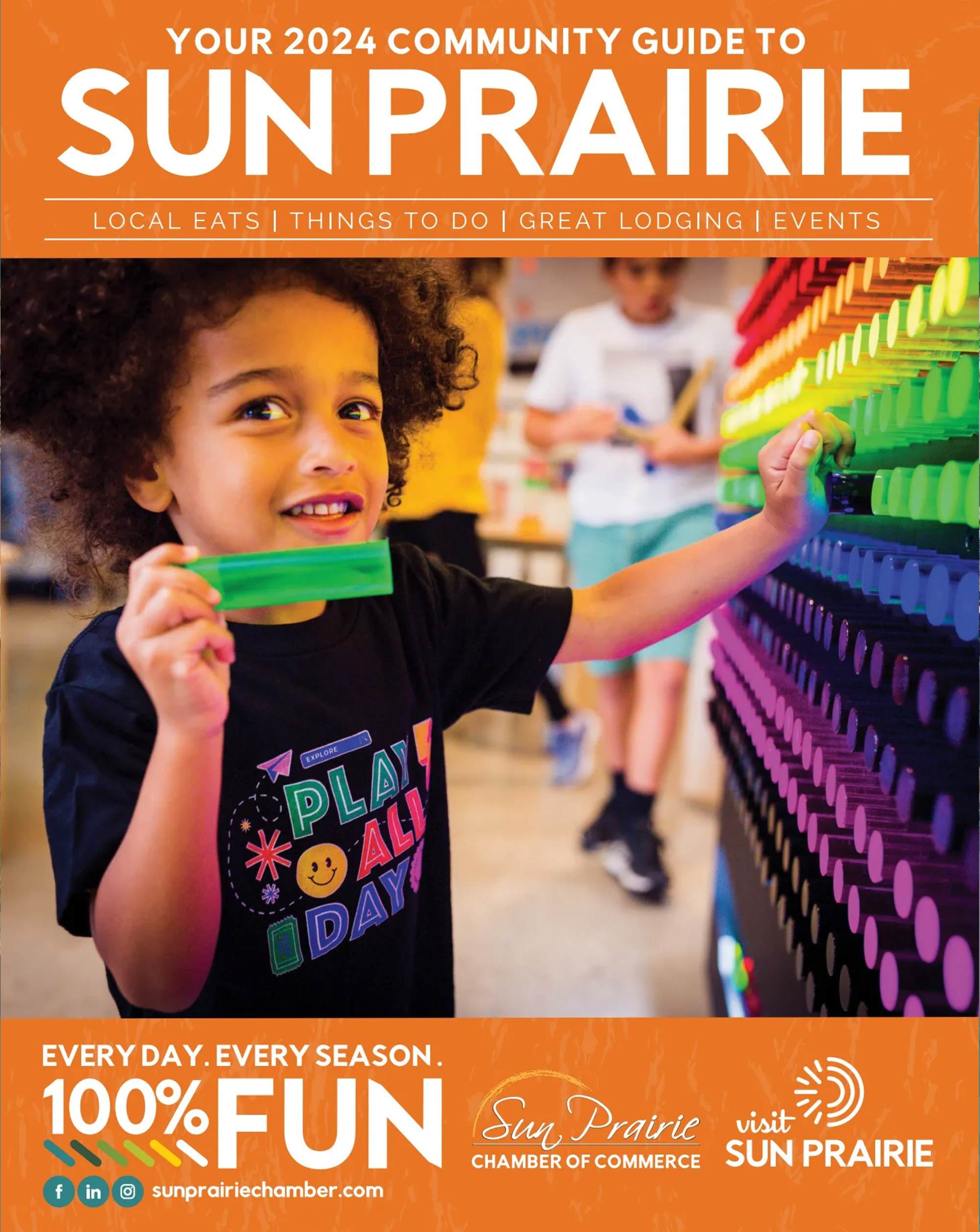 A child reaches for a brightly colored object on a light board at an event. The background features the text, "Your 2024 Community Guide to Sun Prairie" and various logos for Sun Prairie organizations.