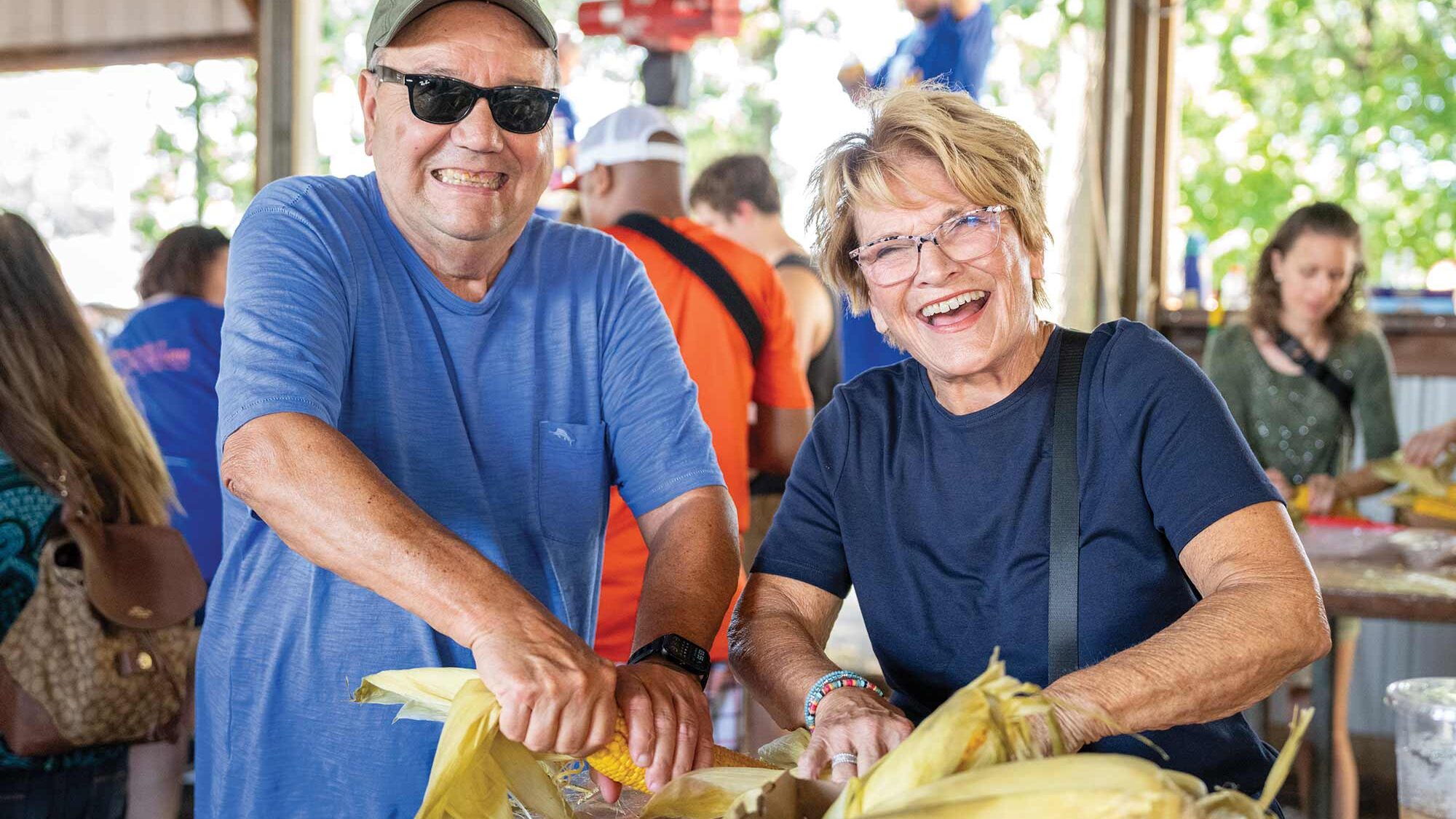 Two smiling people are husking corn at an outdoor event. Others, including one person in the background wearing blue, are also participating. The setting appears lively and engaging.