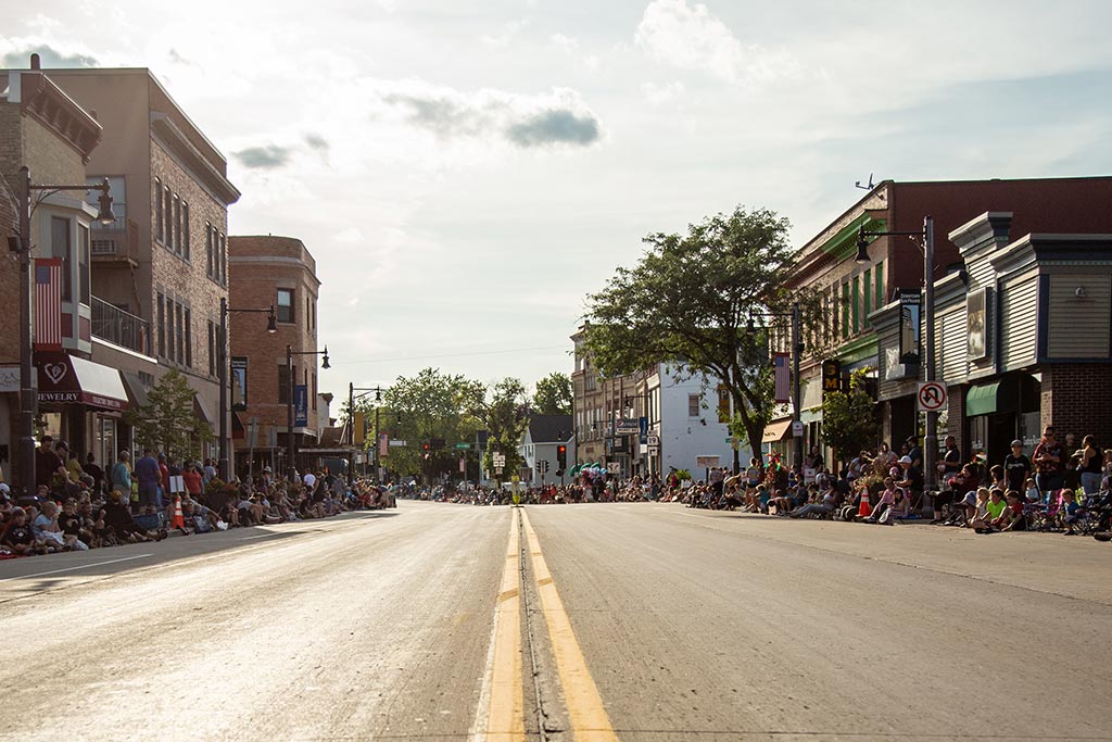 A wide street lined with buildings on both sides is filled with people sitting and standing along the sidewalks, likely waiting for a parade or event to begin. The sky is partly cloudy.