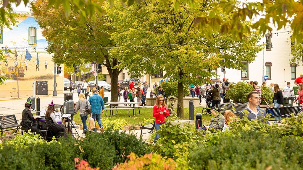 People gathered in a park with trees and benches, engaging in various activities. Some are sitting and talking, while others are walking or standing. Nearby buildings and street scenes are visible.