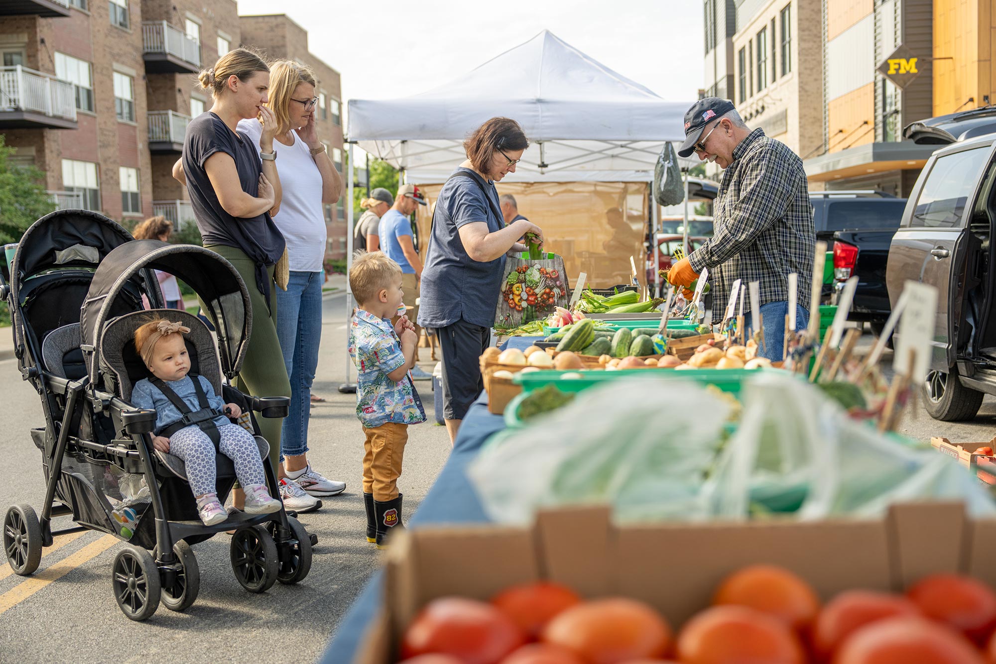 People browse fresh produce at an outdoor market. A woman pushes a stroller with a child, another woman stands nearby, and a man in a hat selects items from a stall.