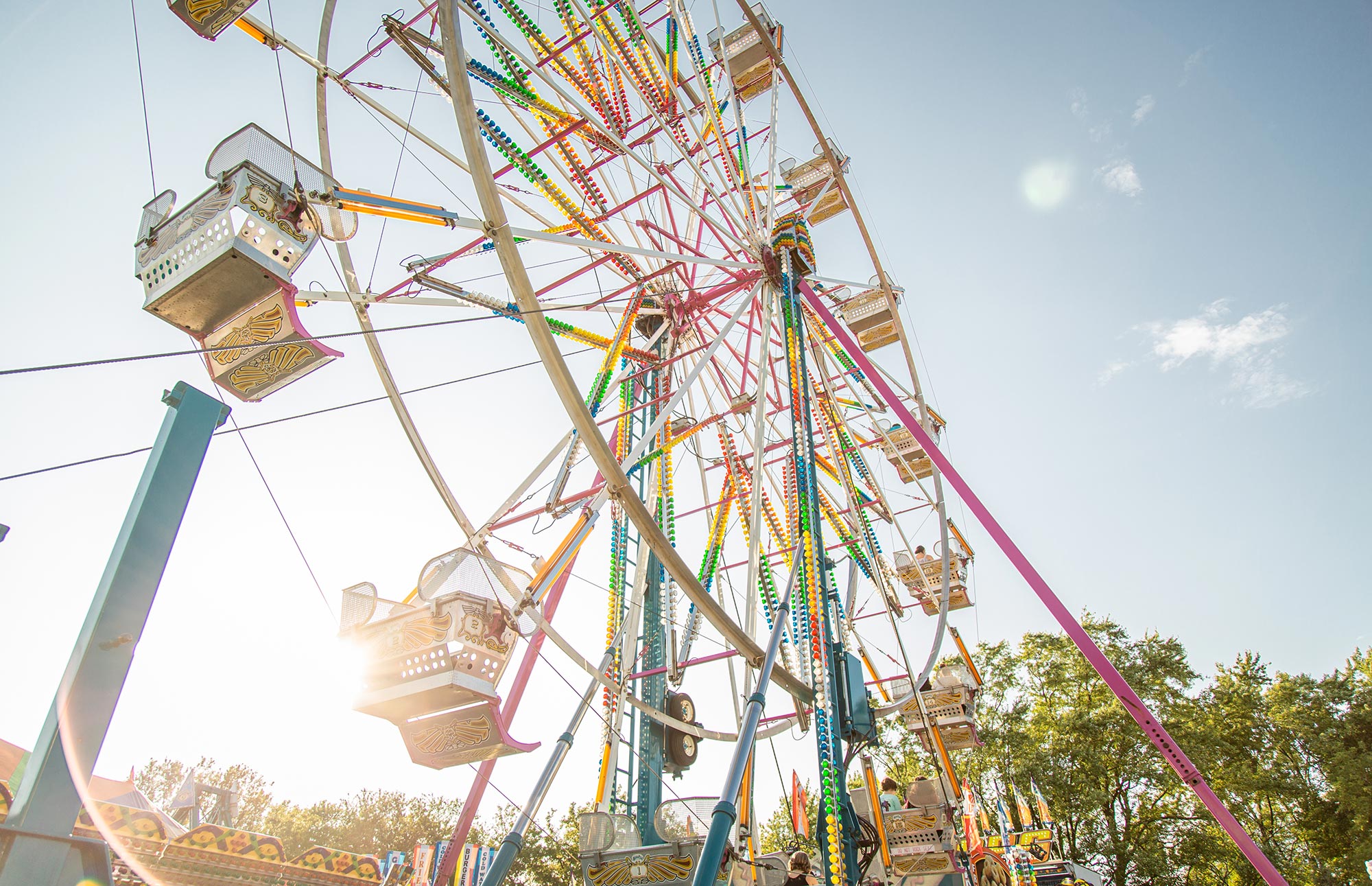 A Ferris wheel against a blue sky, with sunlight shining through its colorful structure. Some trees and parts of other amusement rides are visible in the background.