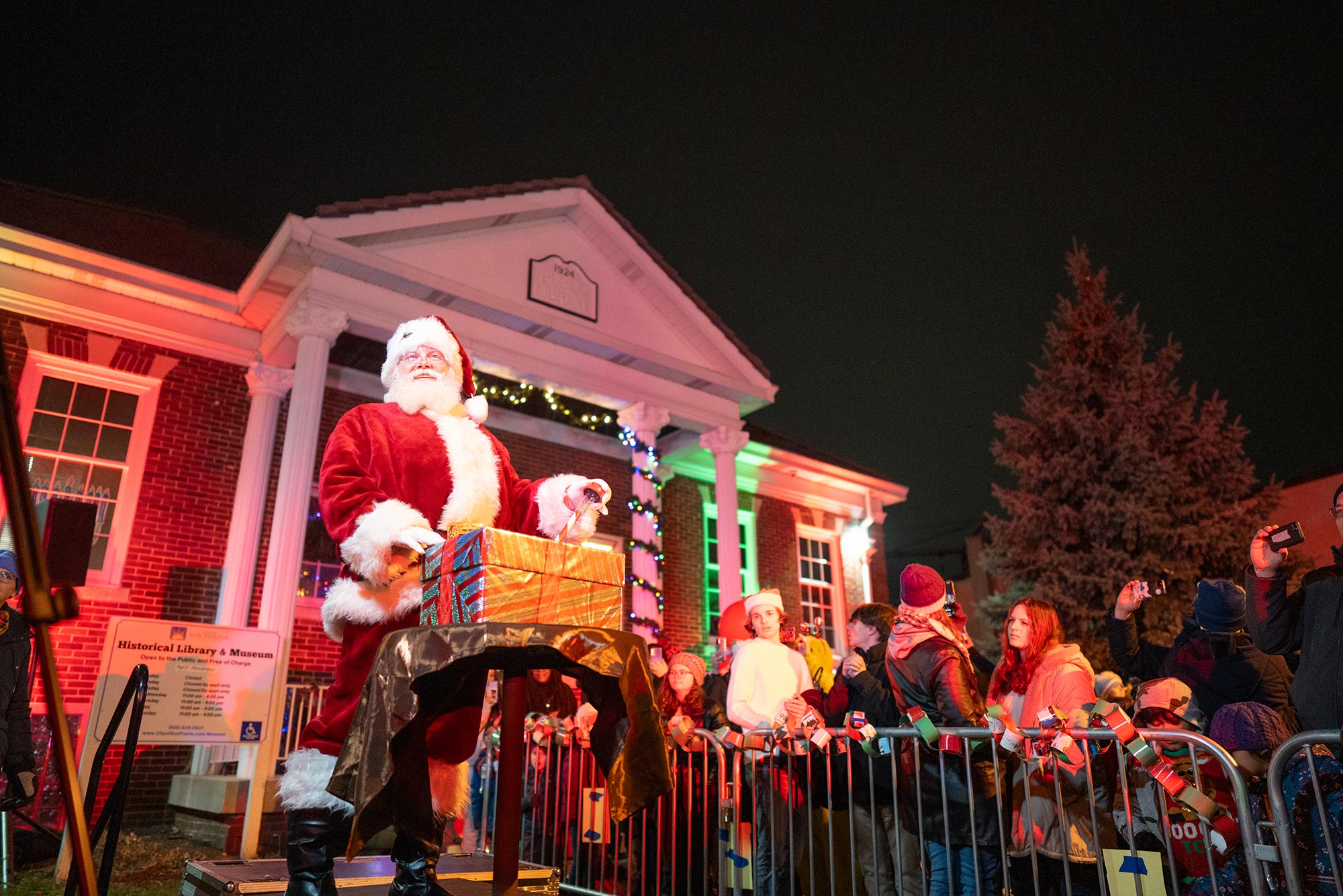 A person dressed as Santa Claus stands on a platform with a dog, surrounded by a festive crowd outside a decorated building at night.