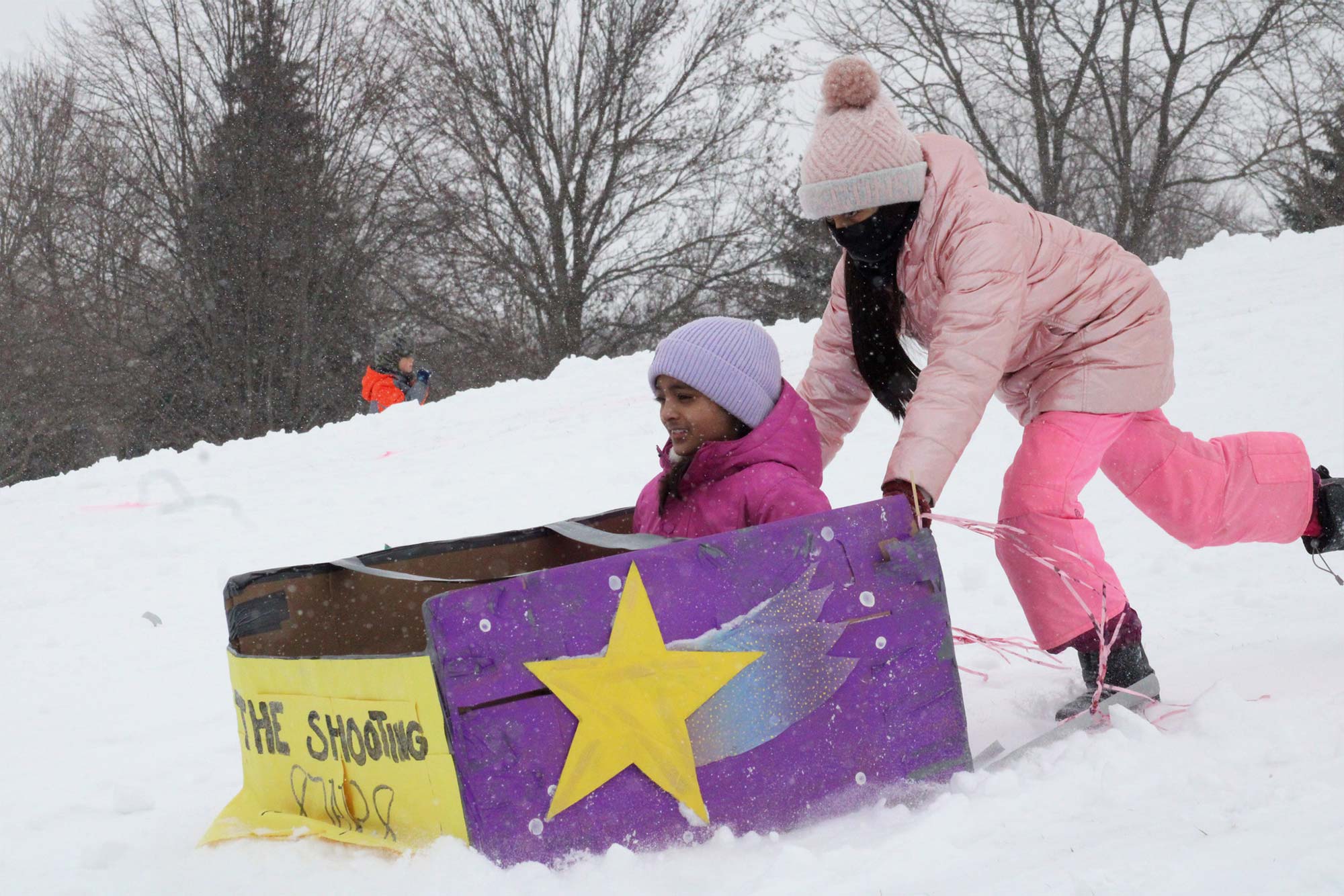 Two children sled down a snowy hill. One child sits in a decorated cardboard sled, while the other stands behind, pushing it. Both are dressed in winter clothing.