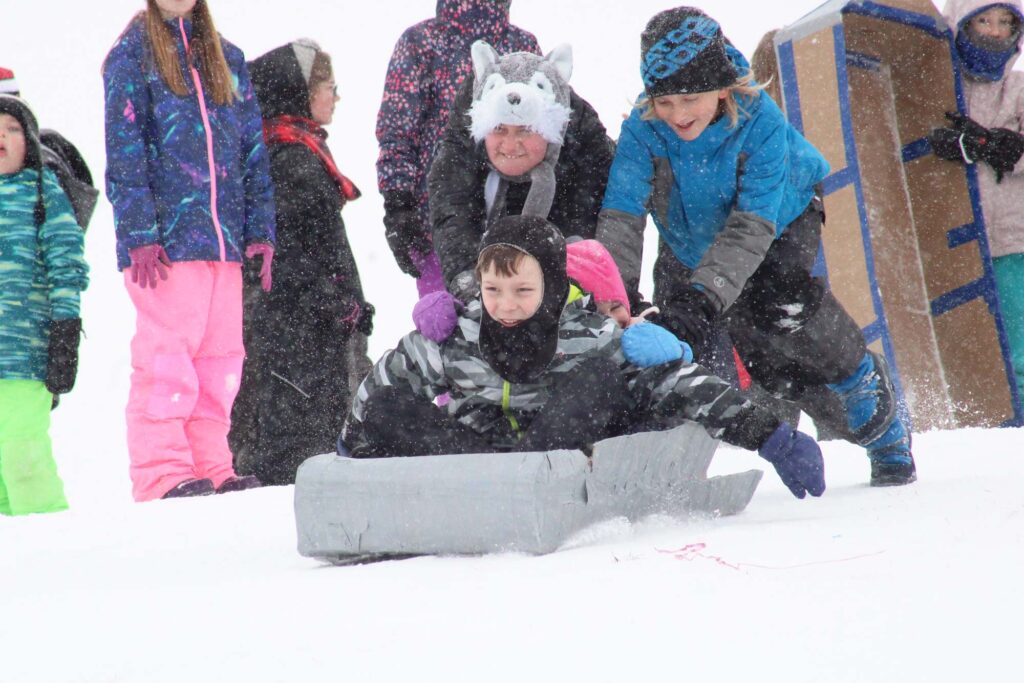 Children, dressed for winter, participate in a sledding race on a snowy hill. One child rides a cardboard sled as others push and cheer. Snow is visibly falling around them.