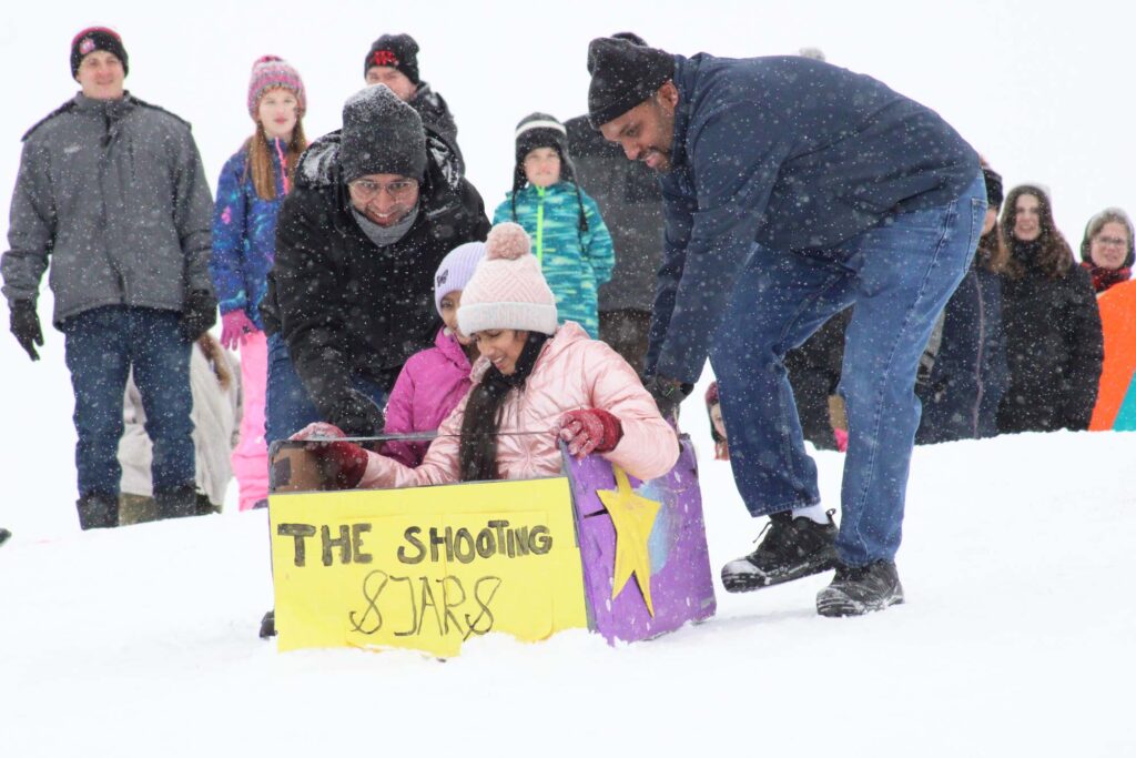 Two adults help two children in a cardboard sled labeled "The Shooting Stars" down a snowy hill, with a crowd of people in winter clothing watching in the background.