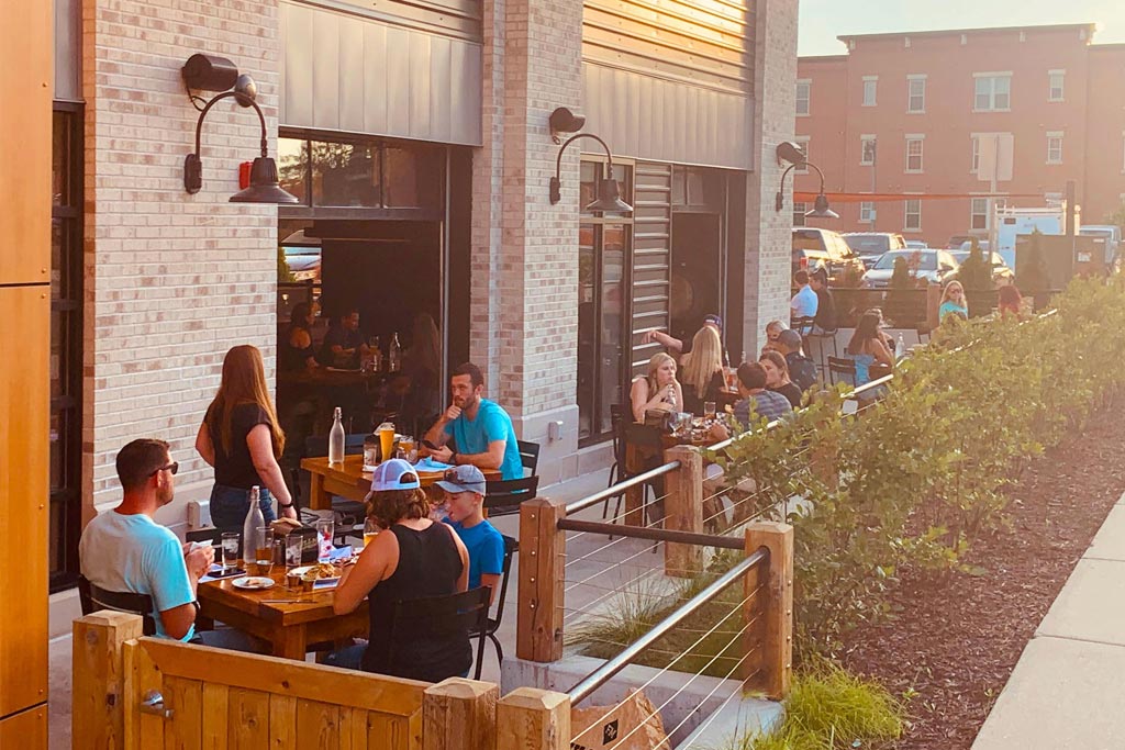 People dining at outdoor tables of a restaurant with a brick exterior. Nearby, other patrons are enjoying their meals and conversations. There is some greenery and a sidewalk in the foreground.