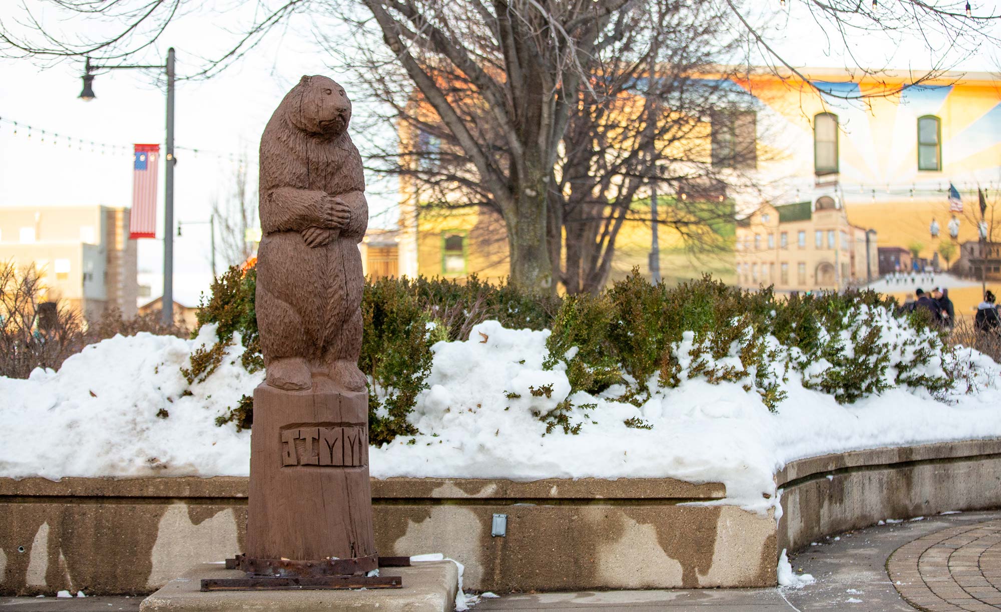 A groundhog statue stands on a pedestal labeled "Jimmy" in a snow-covered park. A mural on a building and barren trees are visible in the background.