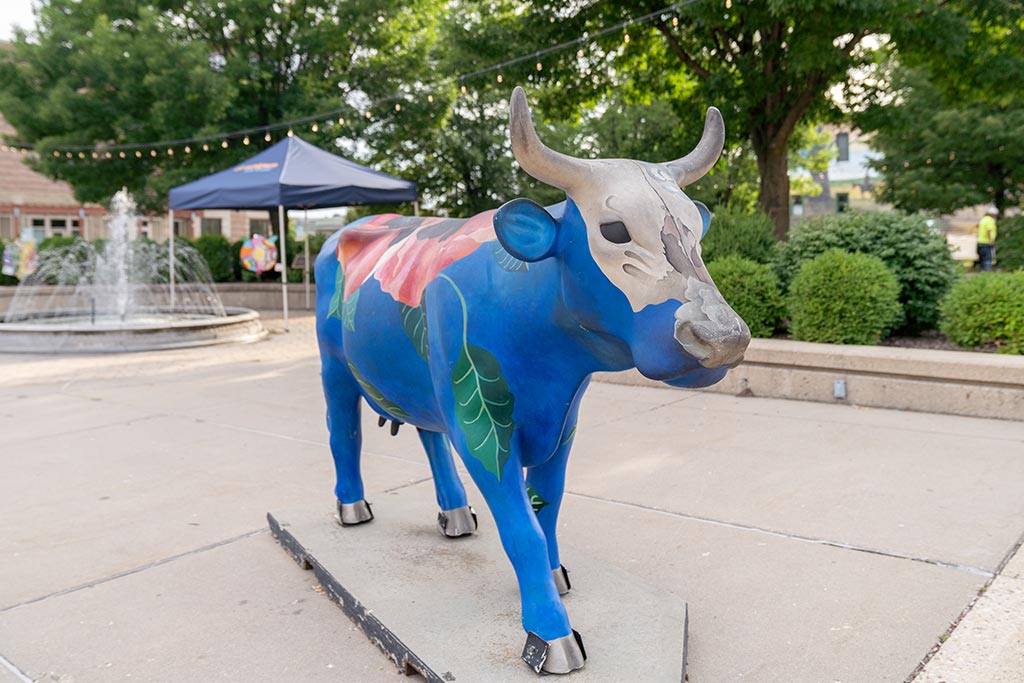 A colorful cow statue, painted with a blue base and large red flowers, is displayed outdoors near a fountain and shaded by nearby trees.