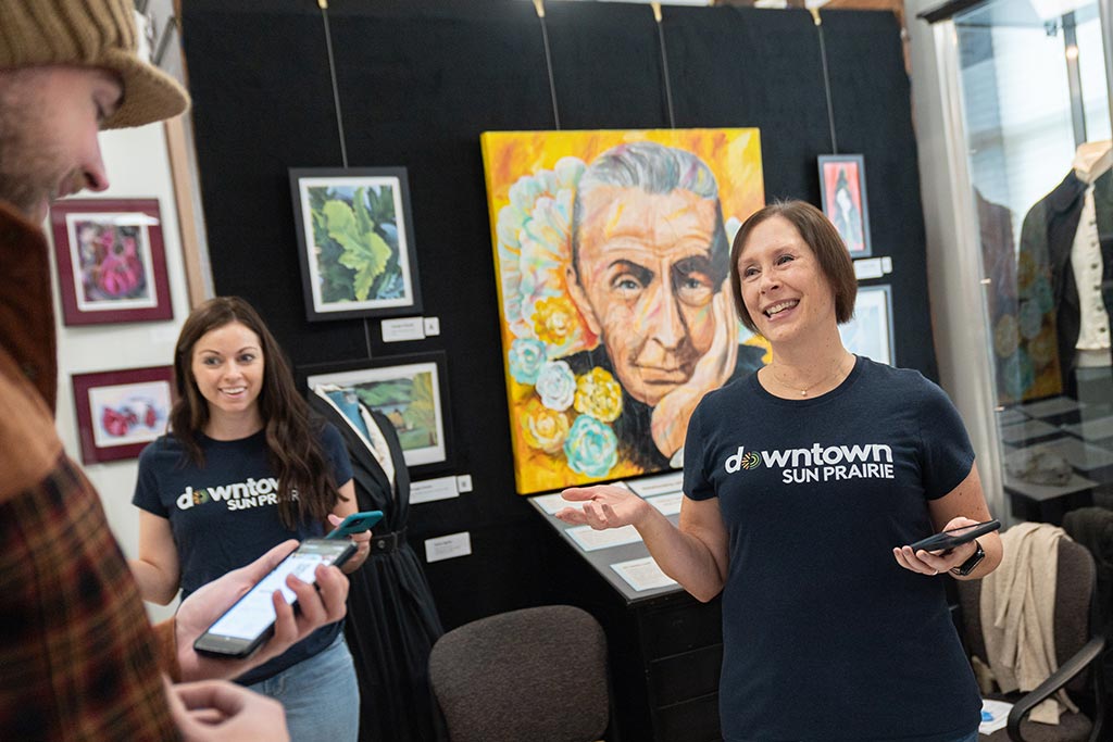 Two women wearing "Downtown Sun Prairie" T-shirts interact with a man. They stand in front of colorful artwork in a gallery setting.