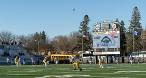 Football players on a field during a game at Bank of Sun Prairie Stadium. A ball is in the air, and the scoreboard in the background displays "Isthmus Bowl." Spectators are seated in the bleachers.