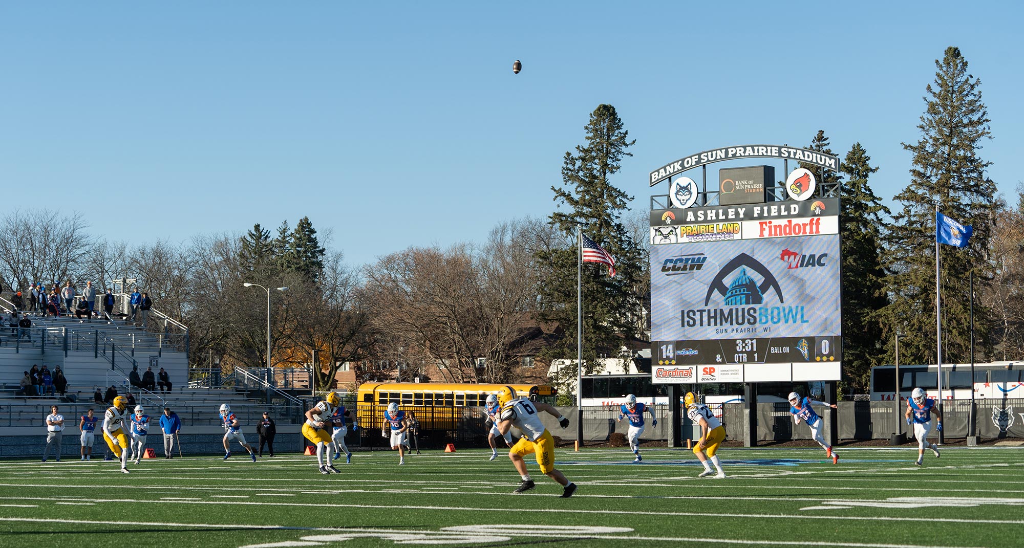 Football players on a field during a game at Bank of Sun Prairie Stadium. A ball is in the air, and the scoreboard in the background displays "Isthmus Bowl." Spectators are seated in the bleachers.