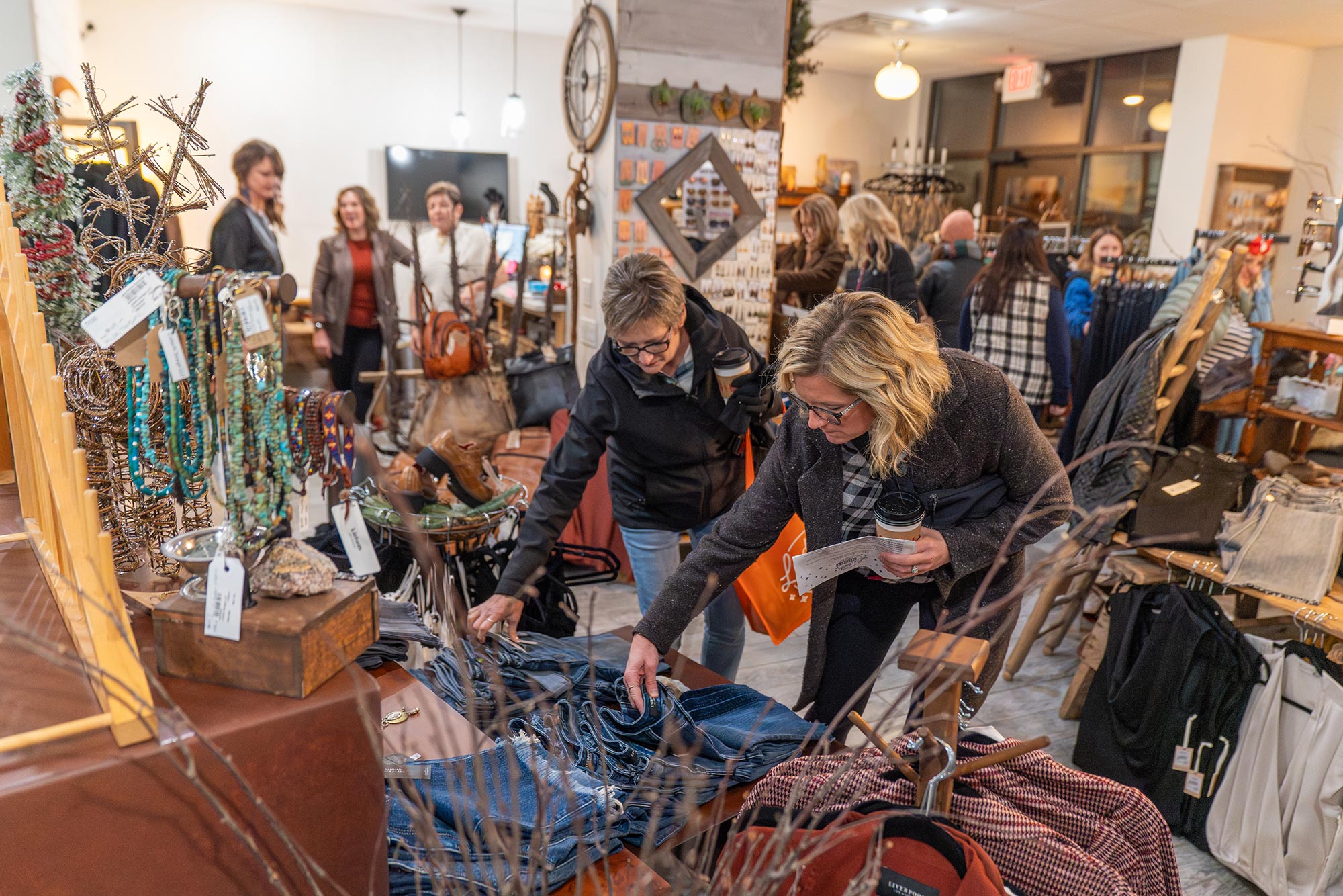 People are shopping and browsing various items in a busy, well-lit store. Two women in the foreground are closely inspecting clothing and accessories displayed on a table.