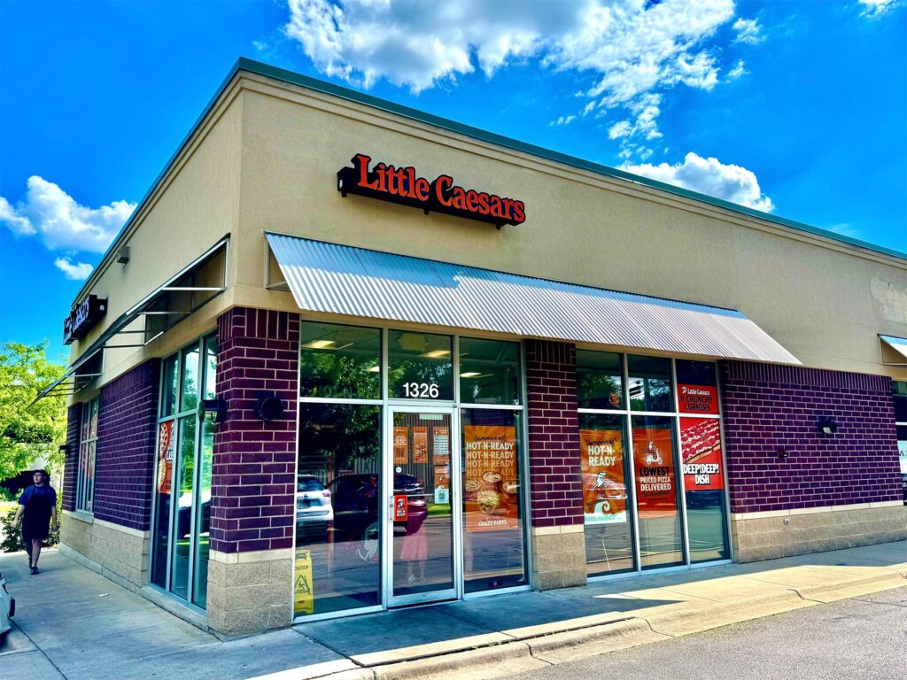 Exterior of a Little Caesars pizza restaurant on a sunny day, located at 1326. The building features glass doors and windows, and a sign for Little Caesars is displayed at the top.