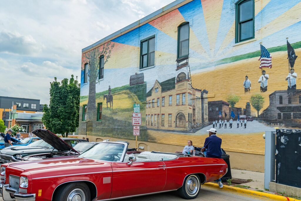 A red convertible car is parked in front of a large mural on a building wall depicting historical scenes. Two people are standing near the car.