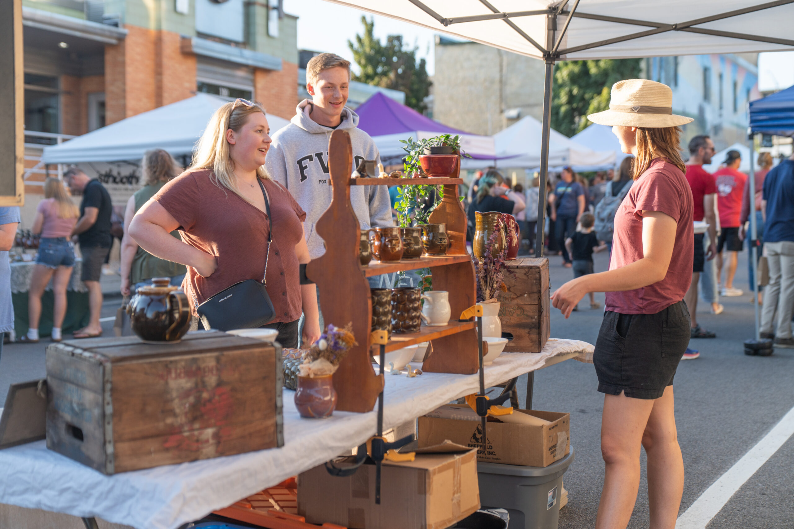 Three people stand at an outdoor market booth with handmade ceramics and wooden items displayed on shelves under a canopy.