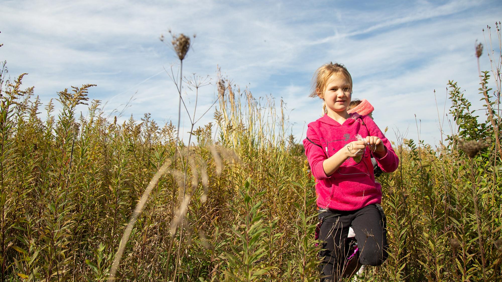 A young girl in a pink hoodie stands in a field of tall grass and plants, holding something in her hands. The sky is clear with scattered clouds.