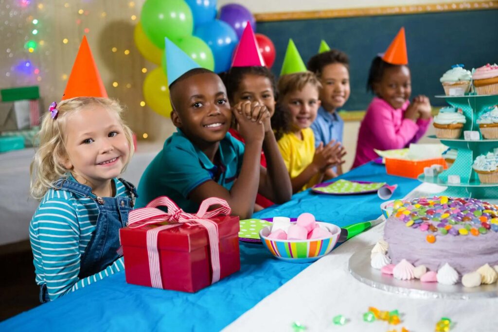 A group of children wearing party hats sit at a table decorated with colorful balloons, enjoying a birthday party with a large cake, cupcakes, and presents.