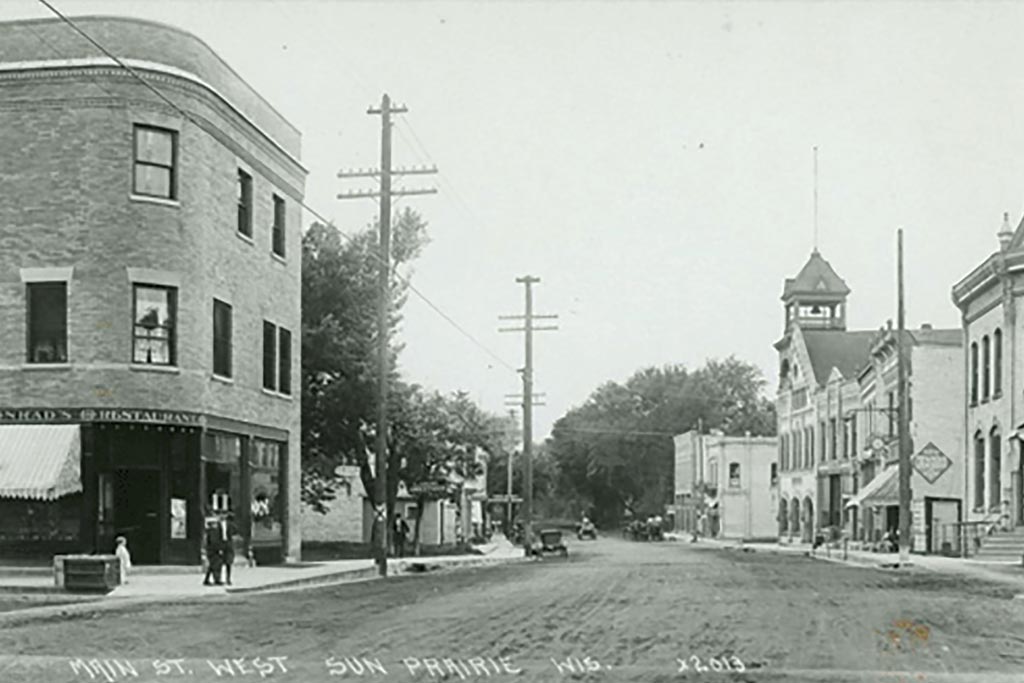 Black and white photo of Main St. West in Sun Prairie, Wisconsin, featuring early 20th-century buildings, people walking, and a few parked cars.