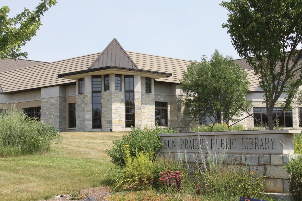 A modern stone building with a triangular roof section and large windows, set amidst landscaped greenery and flowers, marked by a sign reading "Sun Prairie Public Library.