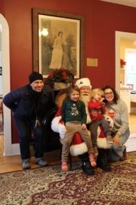 A family poses with Santa Claus inside a warmly decorated room. Two children sit on Santa's lap, while two adults stand beside them, smiling.