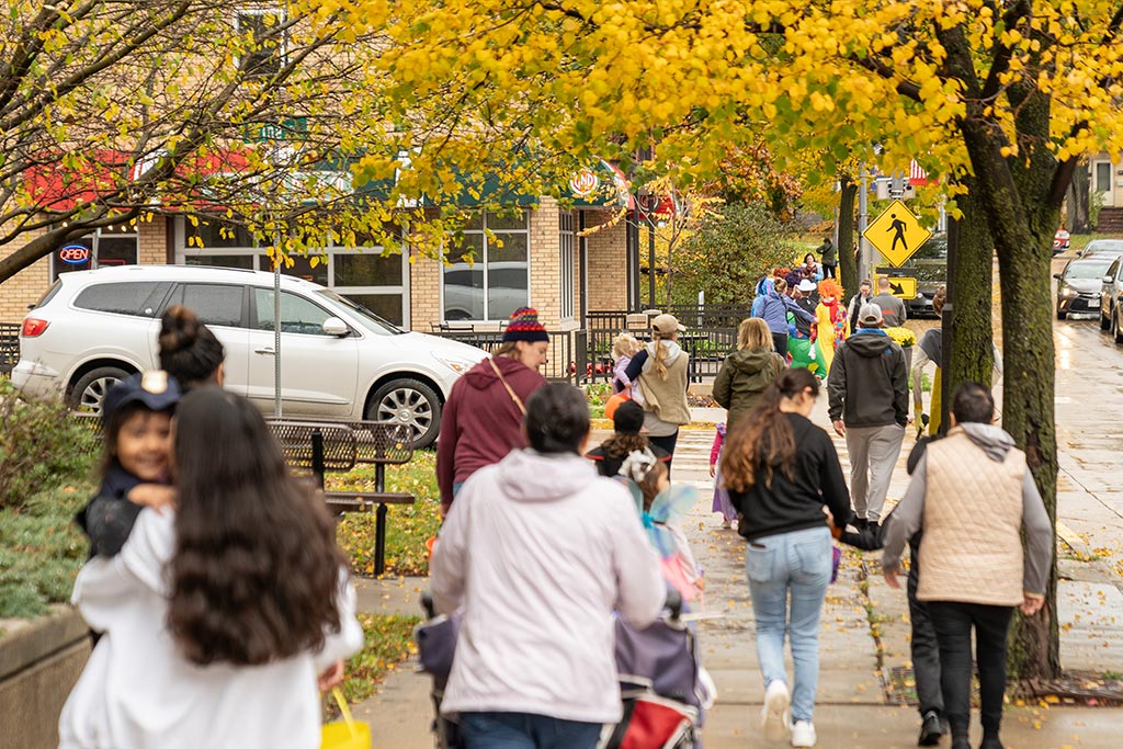 Crowd of people walking along a tree-lined sidewalk, some carrying children. The scene features autumn foliage, a parked car, and a pedestrian crossing sign in the background.