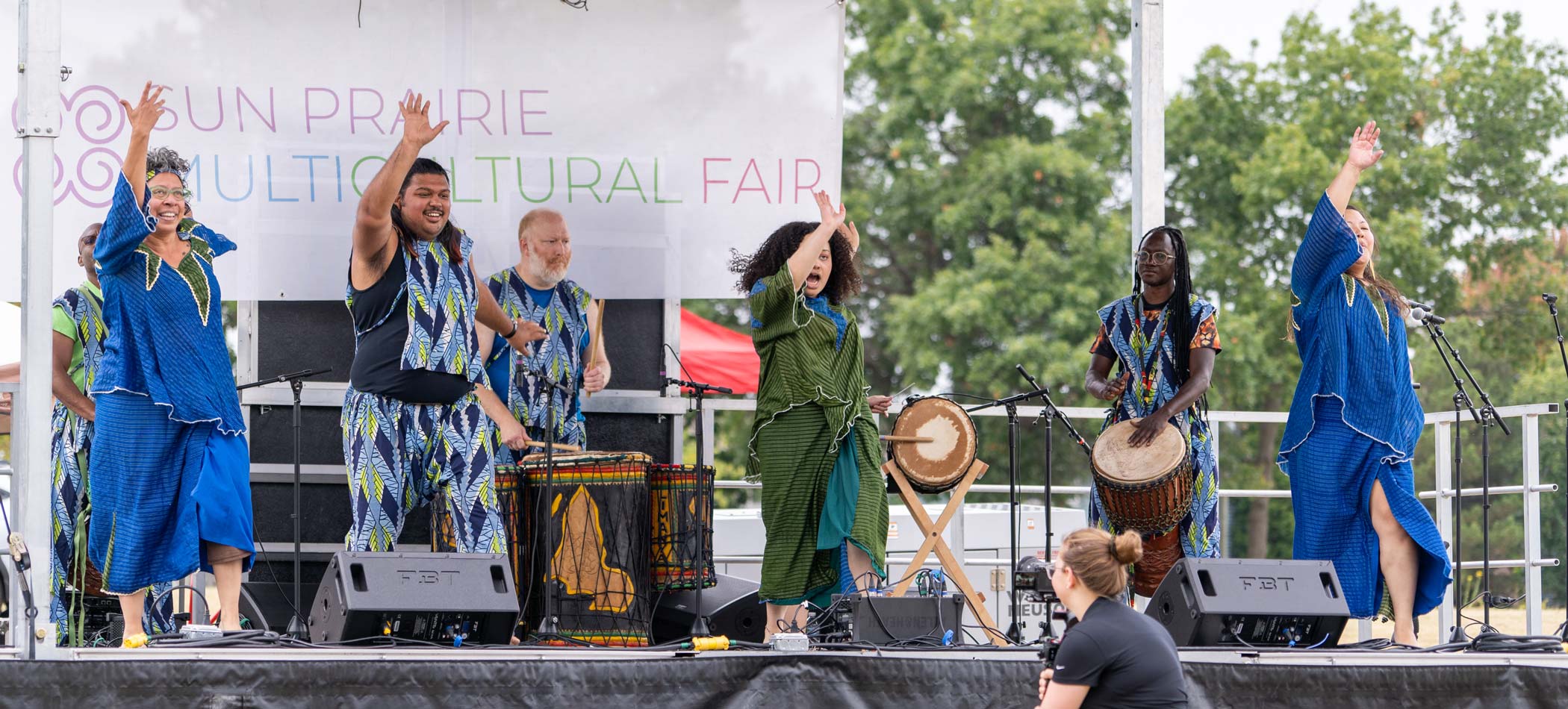 A group of performers in colorful costumes dance and play drums on an outdoor stage during the Sun Prairie Multicultural Fair. An audience member watches them.