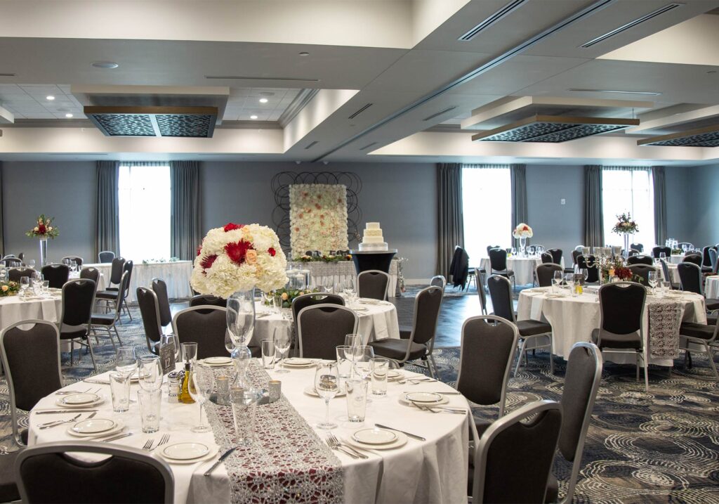 A decorated event space with round tables set with white linens, glassware, and floral centerpieces. A cake stands on a table at the far end of the room surrounded by floral arrangements.