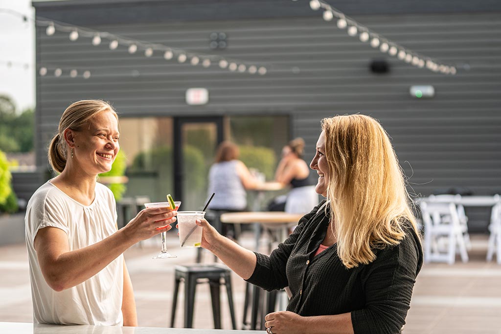 Two women smile and clink glasses at an outdoor bar with string lights overhead. Other patrons can be seen seated in the background.