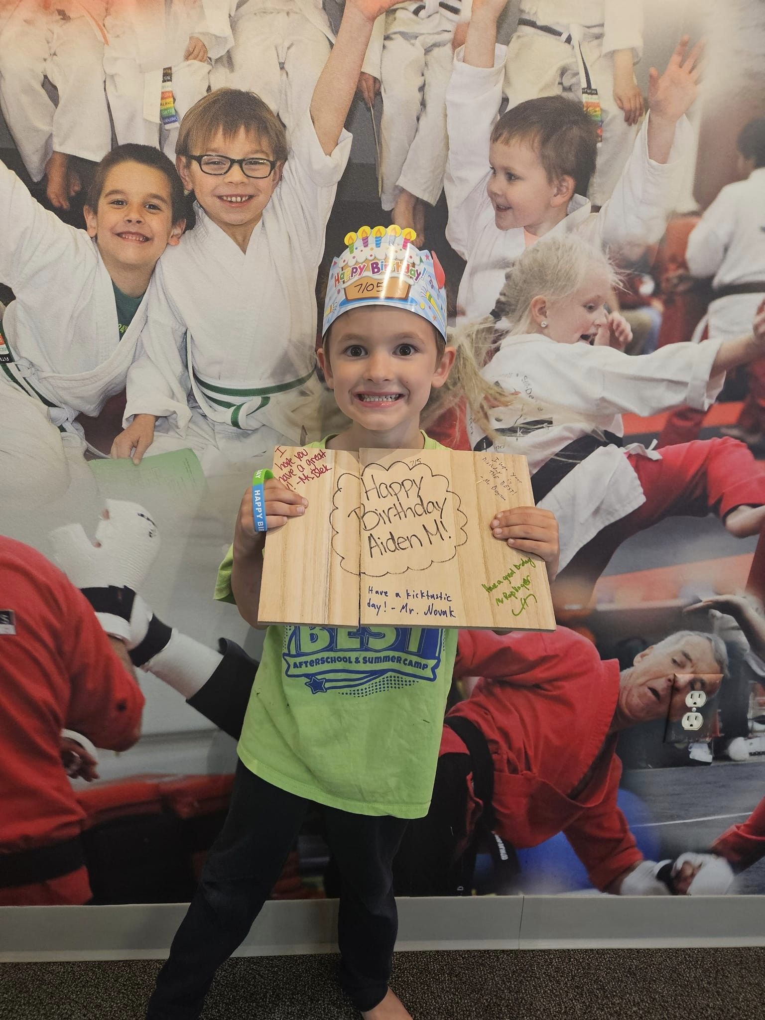 A child wearing a birthday hat and holding a wooden birthday card stands in front of a wall decorated with pictures of children practicing martial arts.