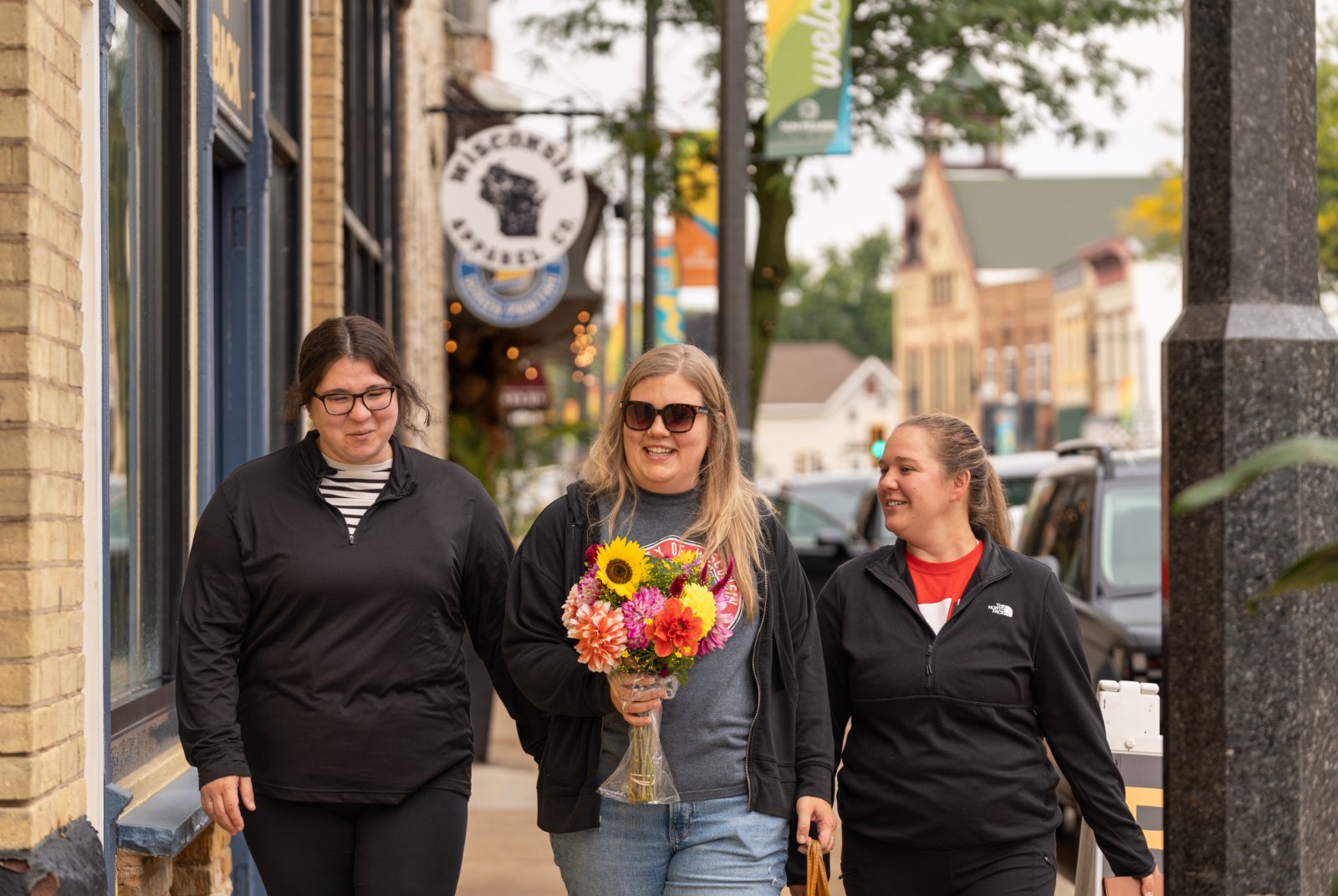 Three women walk on a sidewalk in a small town; one holds a bouquet of flowers. Brick buildings and parked cars line the street.