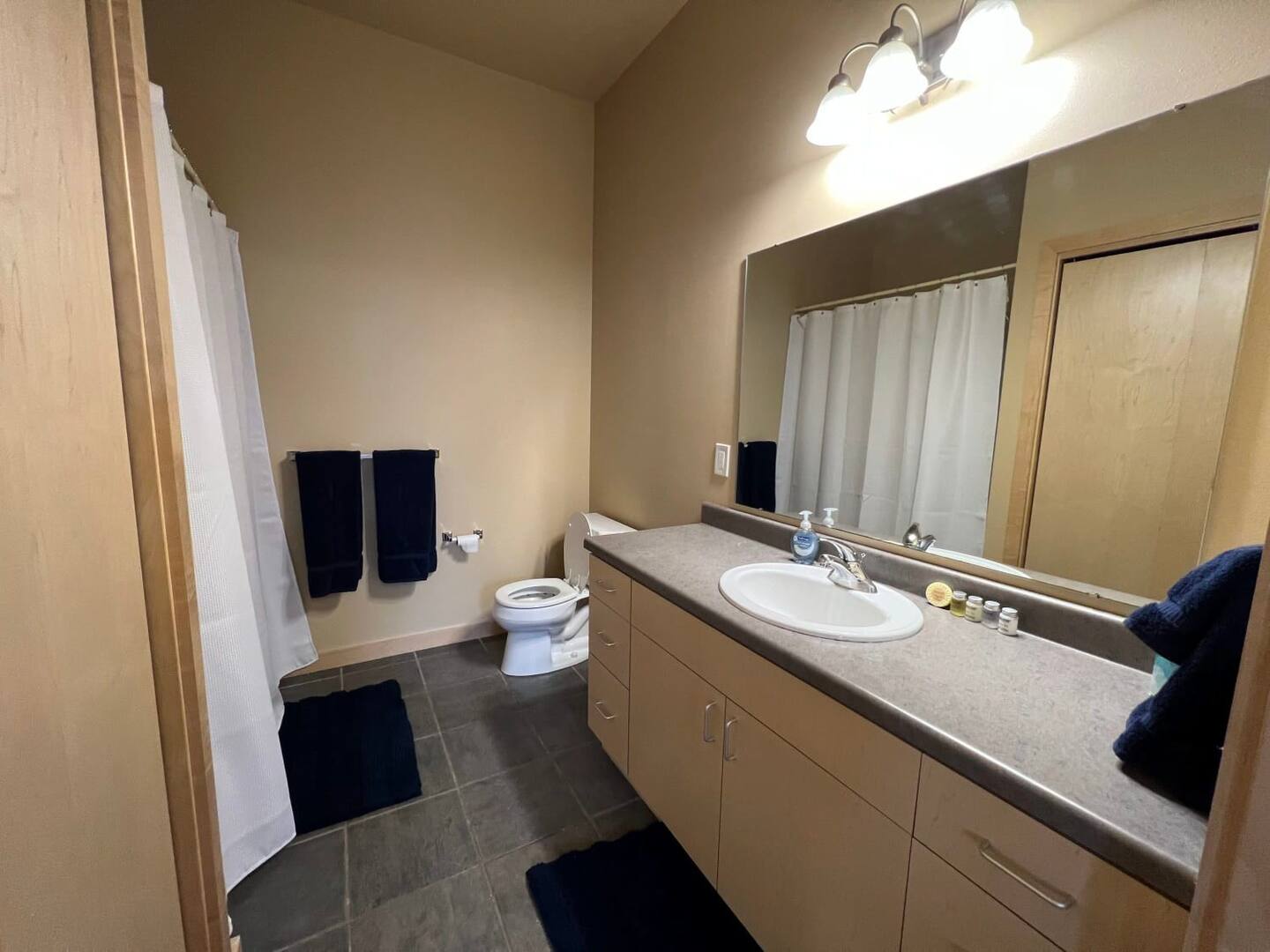 A clean bathroom with a vanity mirror, sink, countertop, toilet, two dark blue towels on the wall, and a shower curtain. The room has beige walls and flooring with dark tiles.