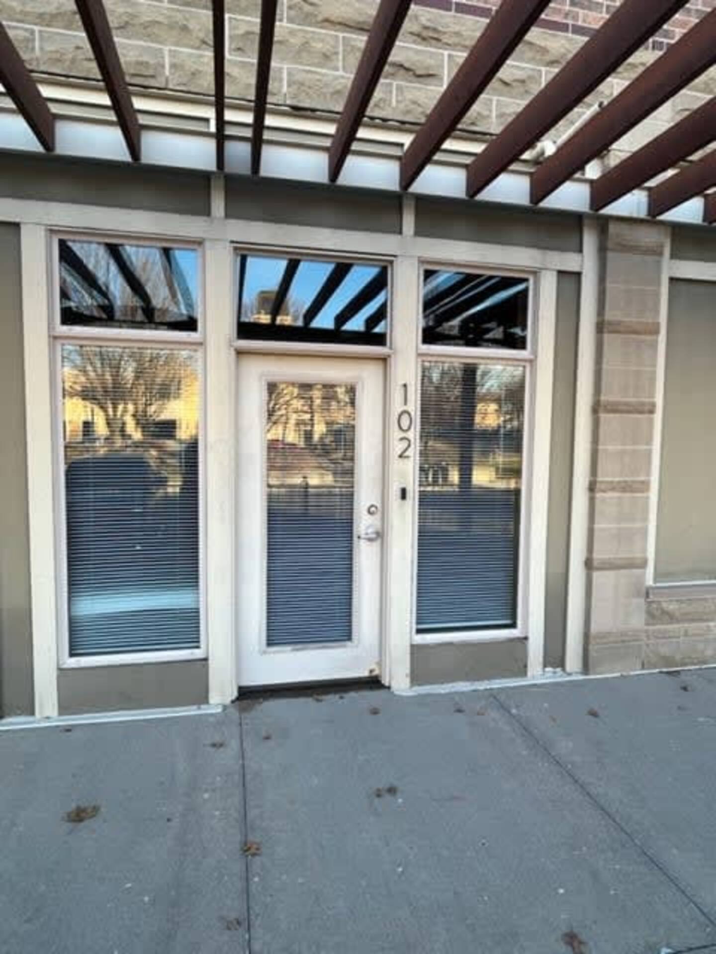 A glass door with the number "102" next to it, flanked by two large windows with closed blinds, set in a stone and concrete building. Reflection of trees visible in the glass.