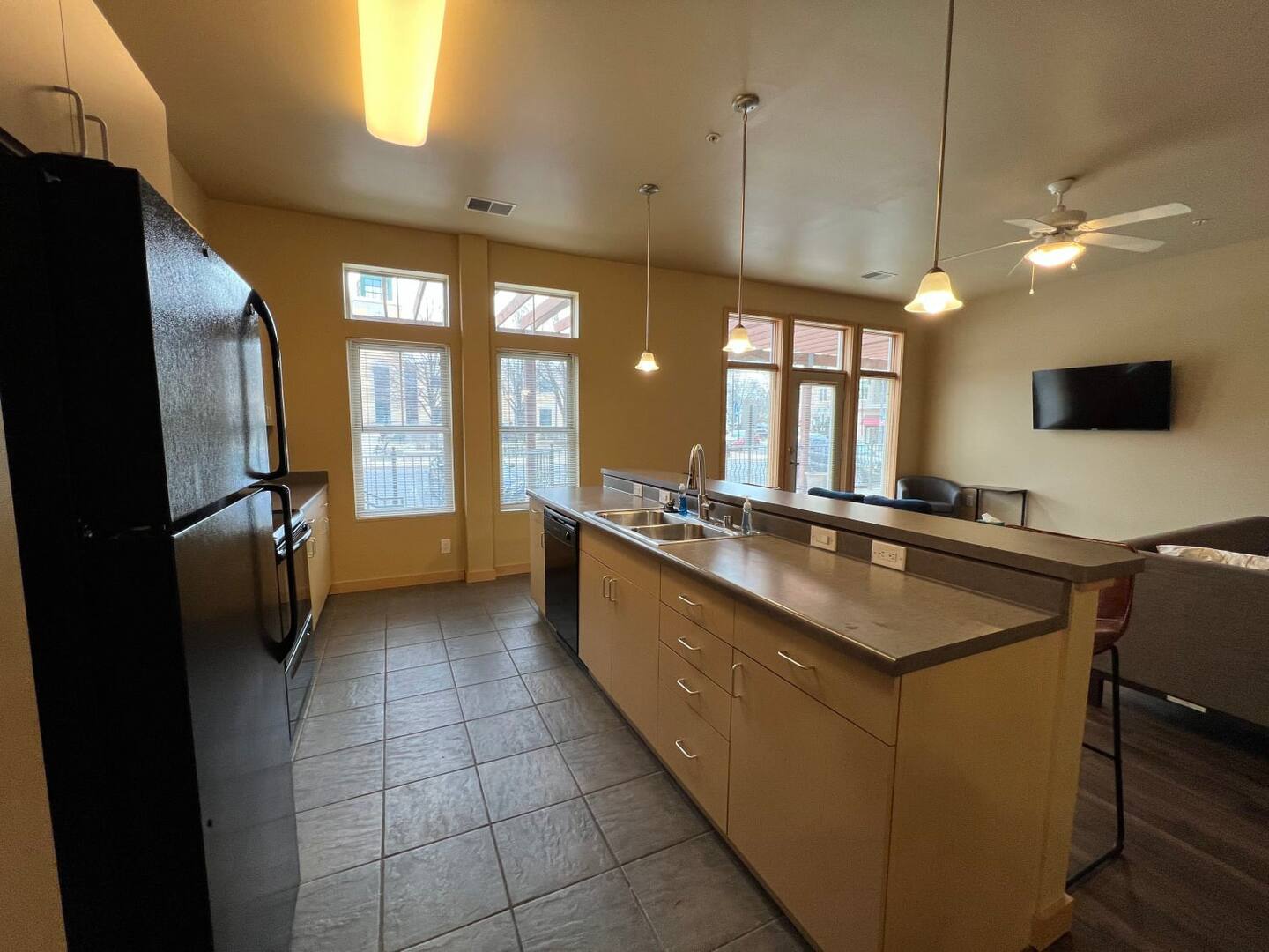 Modern kitchen with tiled floor, light-colored cabinets, and a large island with a sink and bar seating. The space opens into a living area with windows, ceiling lighting, a ceiling fan, and a mounted TV.