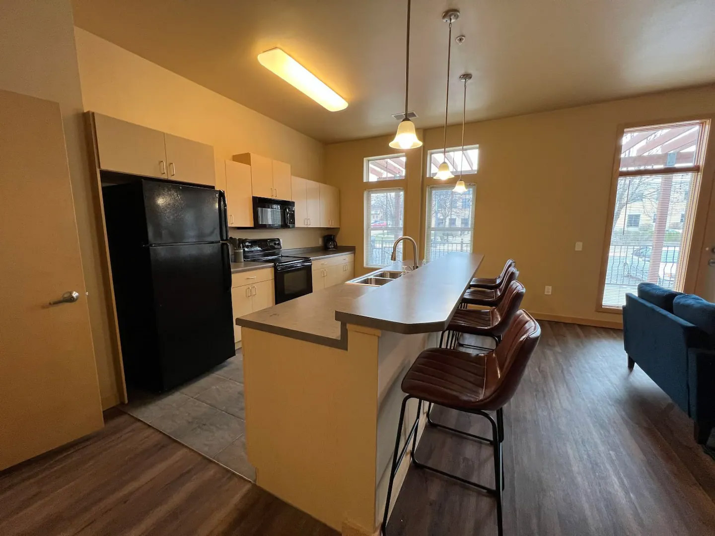 A kitchen and living area with a central island, bar stools, black appliances, beige cabinets, wooden flooring, and pendant lights. Large windows allow natural light to enter the room.