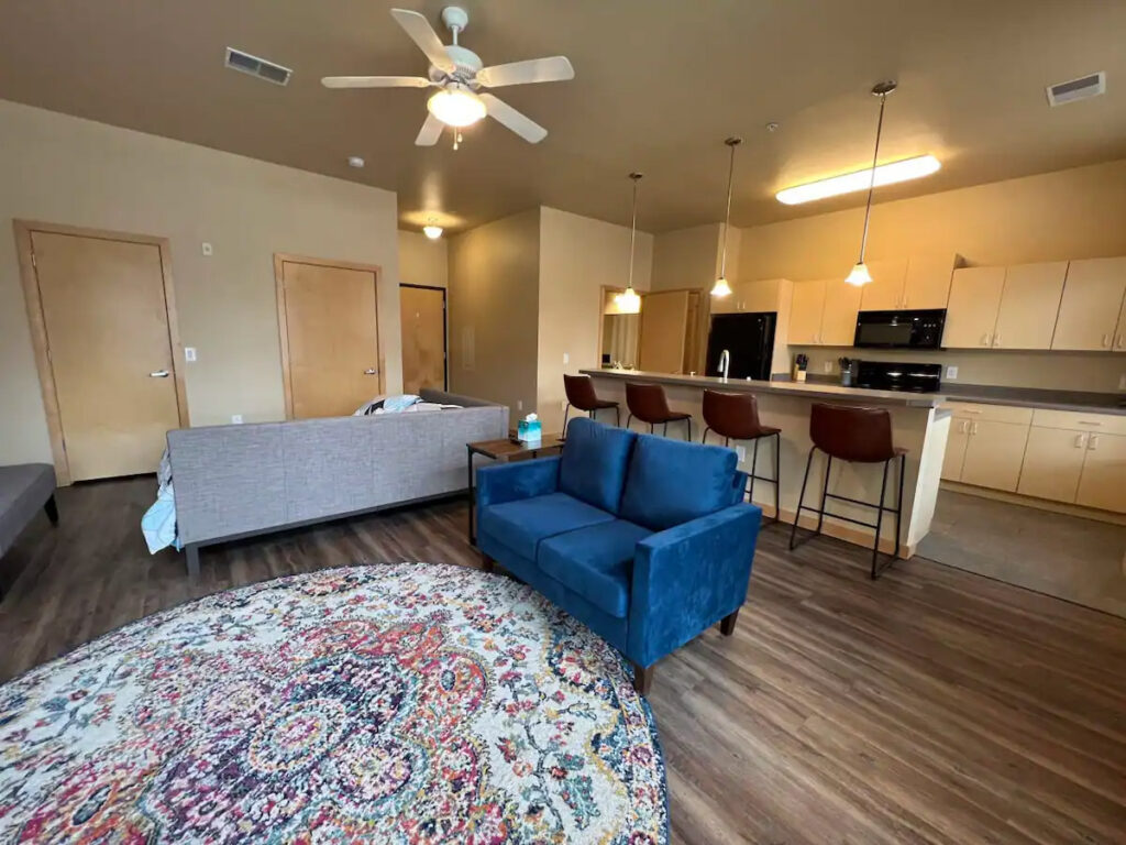 A modern living room with a sofa, blue armchair, patterned rug, ceiling fan, and open kitchen area with bar stools.