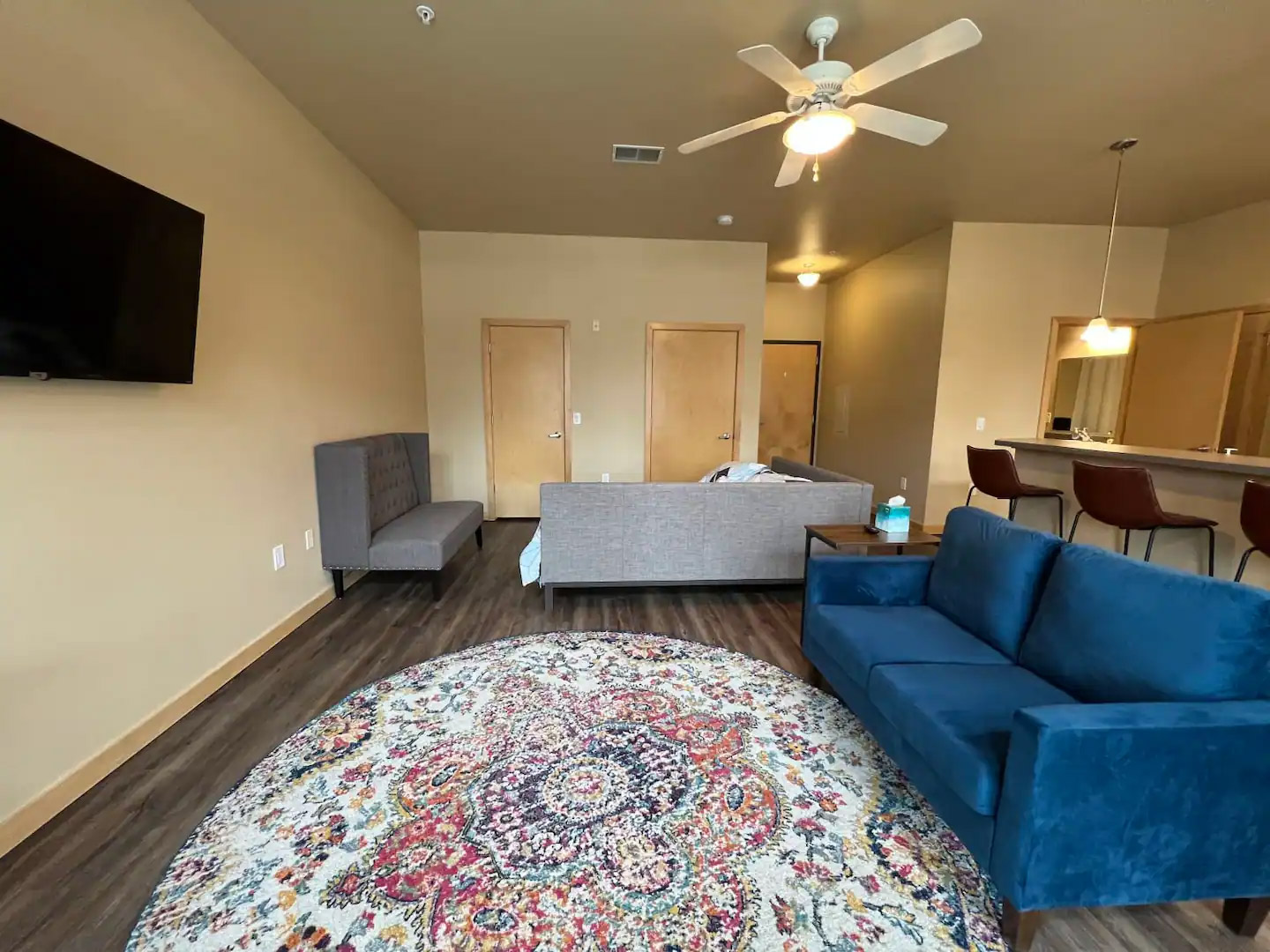 A living room with a blue sofa, gray couch, wall-mounted TV, ceiling fan, and cushioned bench. A round patterned rug covers the floor. A kitchen with bar seating is visible in the background.